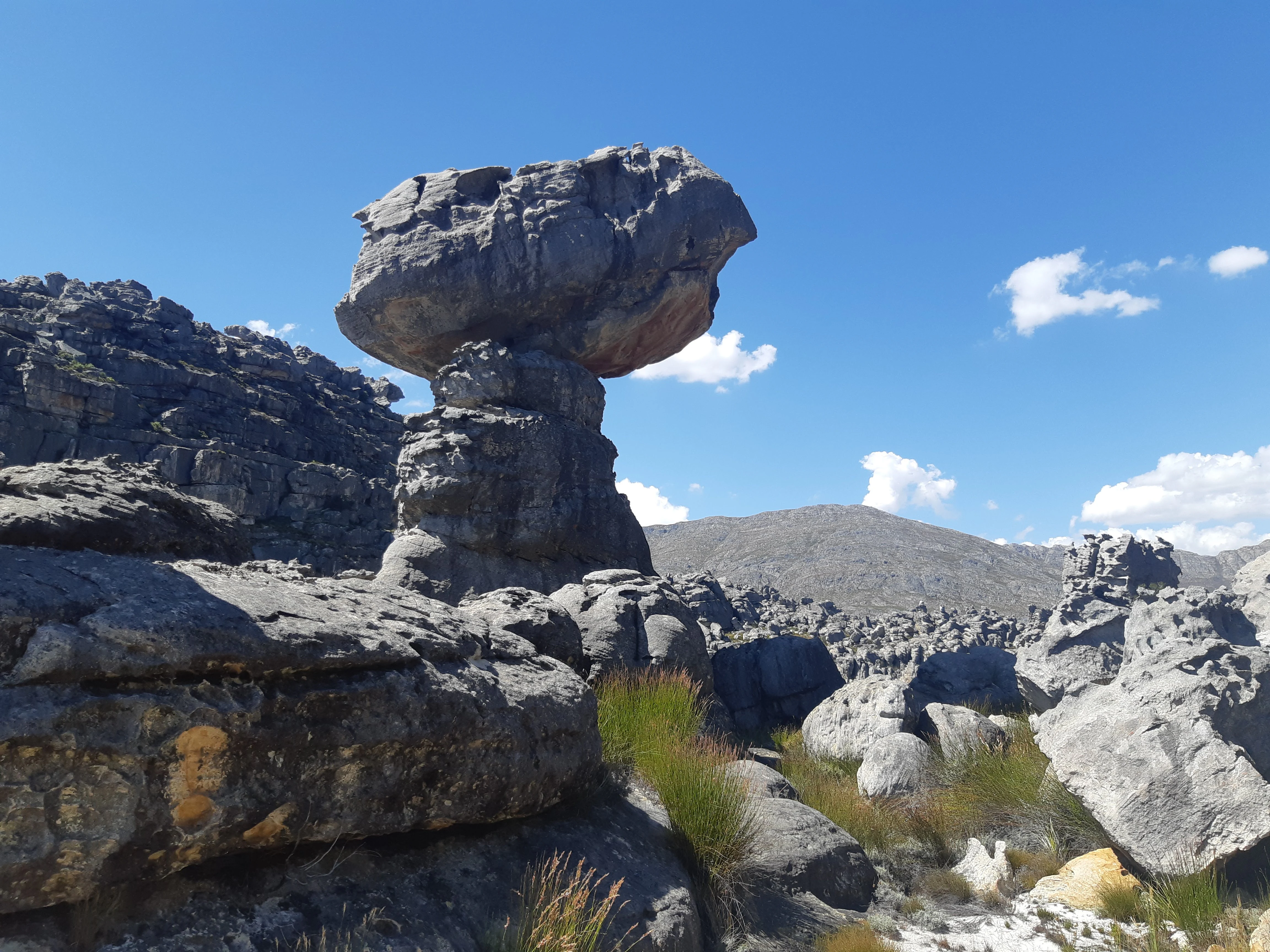A large, weathered sandstone boulder balanced precariously on a smaller rock pedestal under a clear blue sky.
