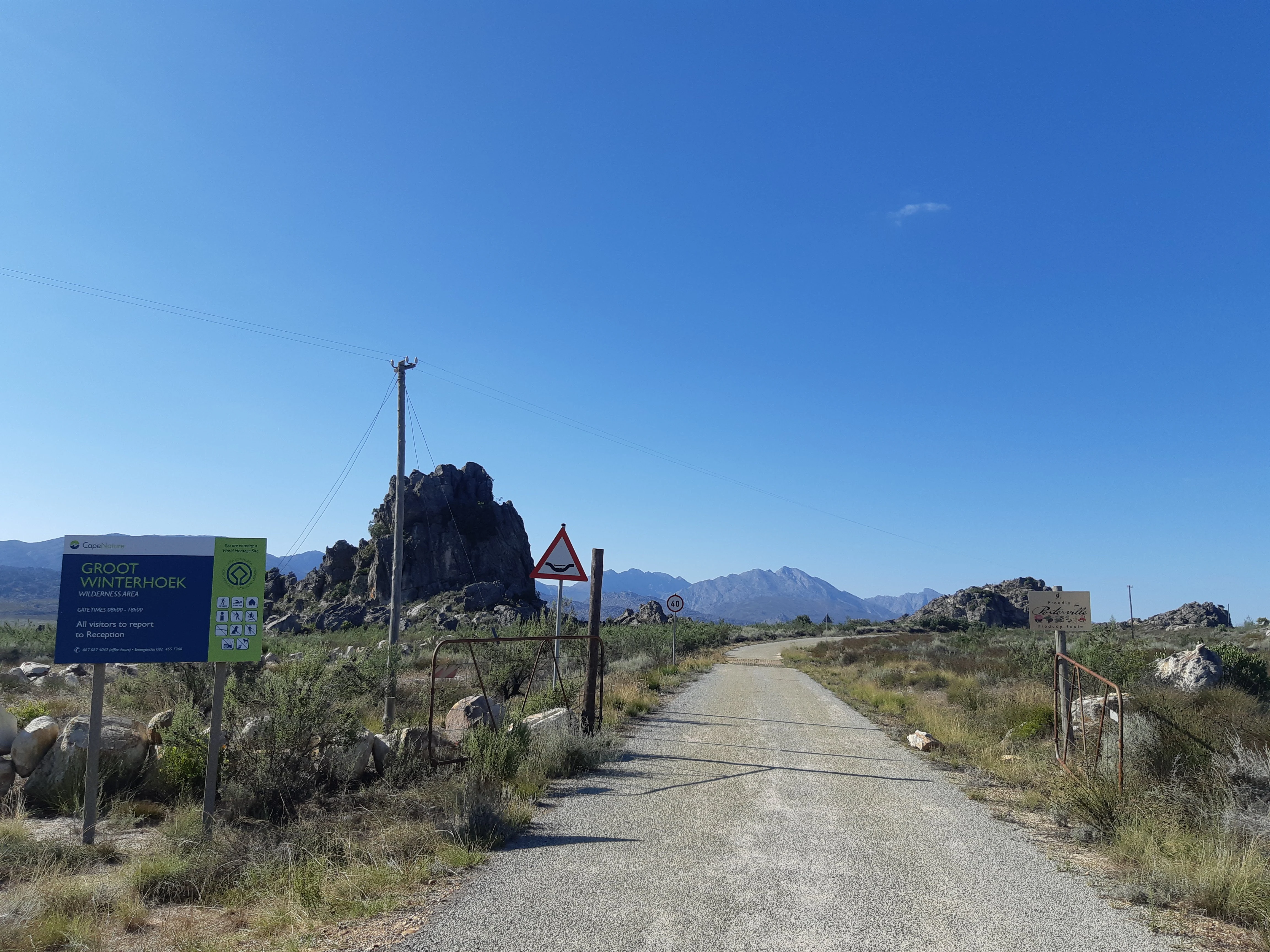 A gravel road leads into a rugged landscape with rocky outcrops under a clear blue sky, featuring a park entrance sign on the left.