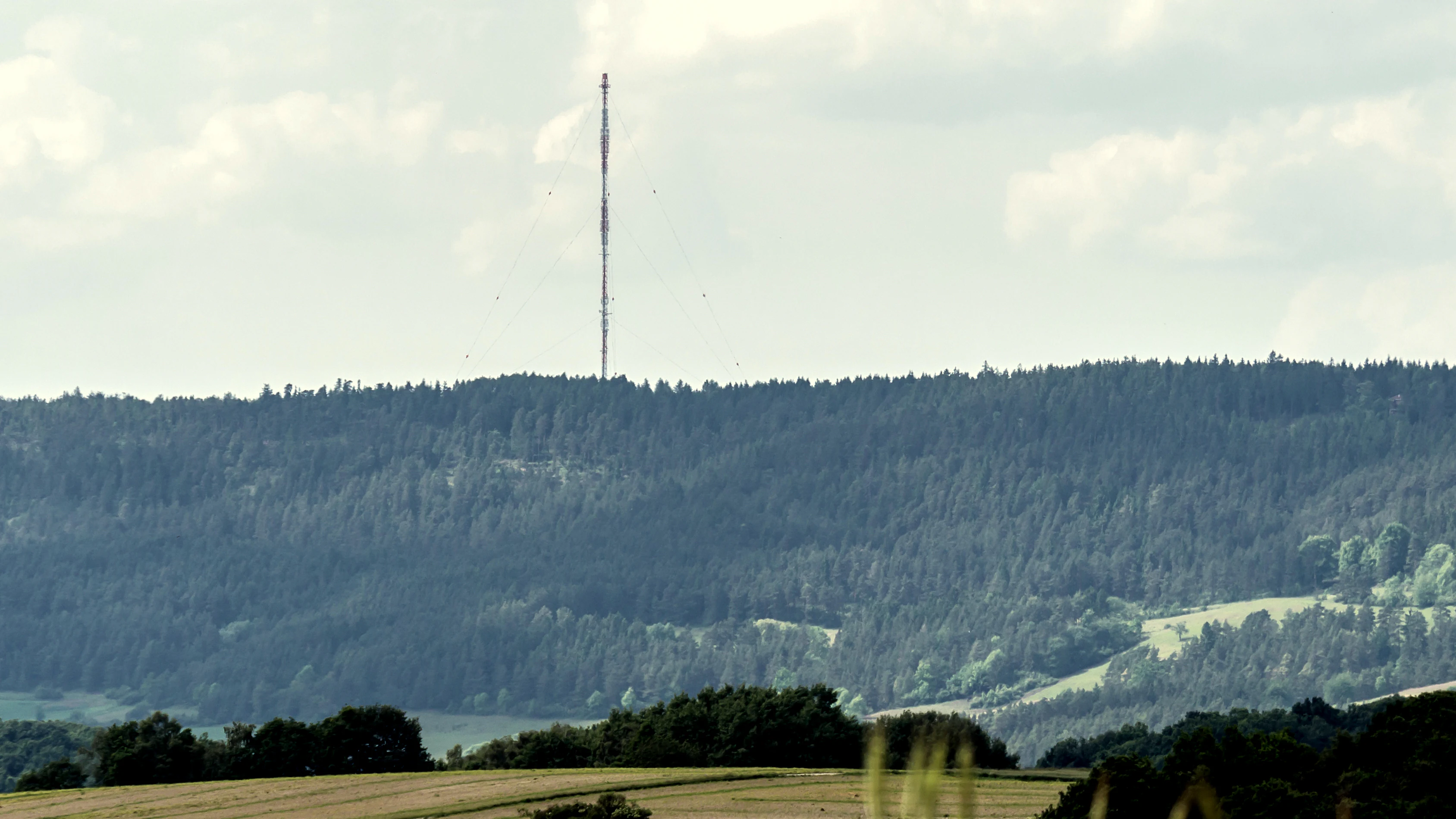 A distant view of a forested mountain slope topped with a tall, thin radio transmission mast under a cloudy sky.