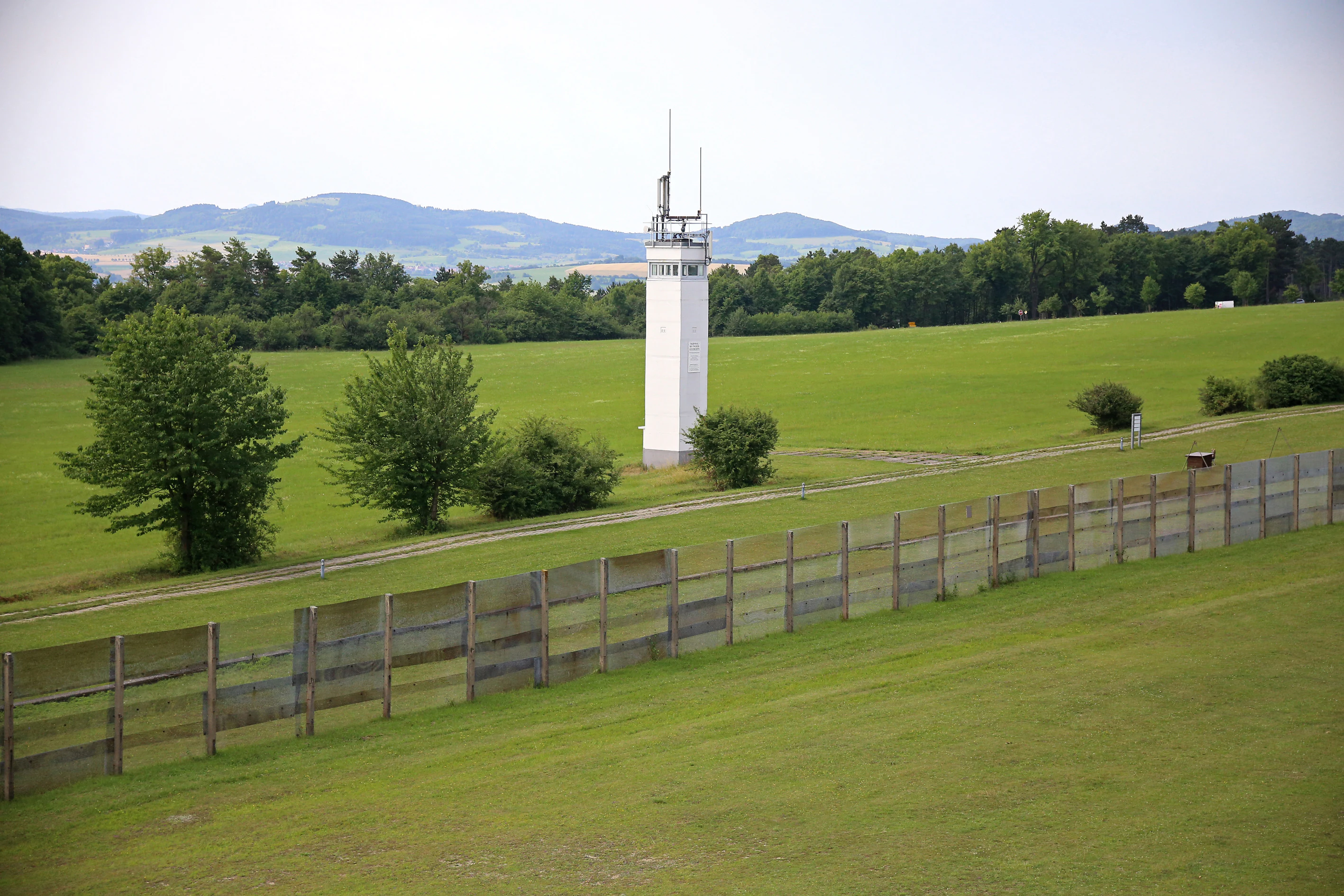 A white observation tower stands in a wide, grassy field alongside a tall metal mesh fence, with rolling hills in the background.