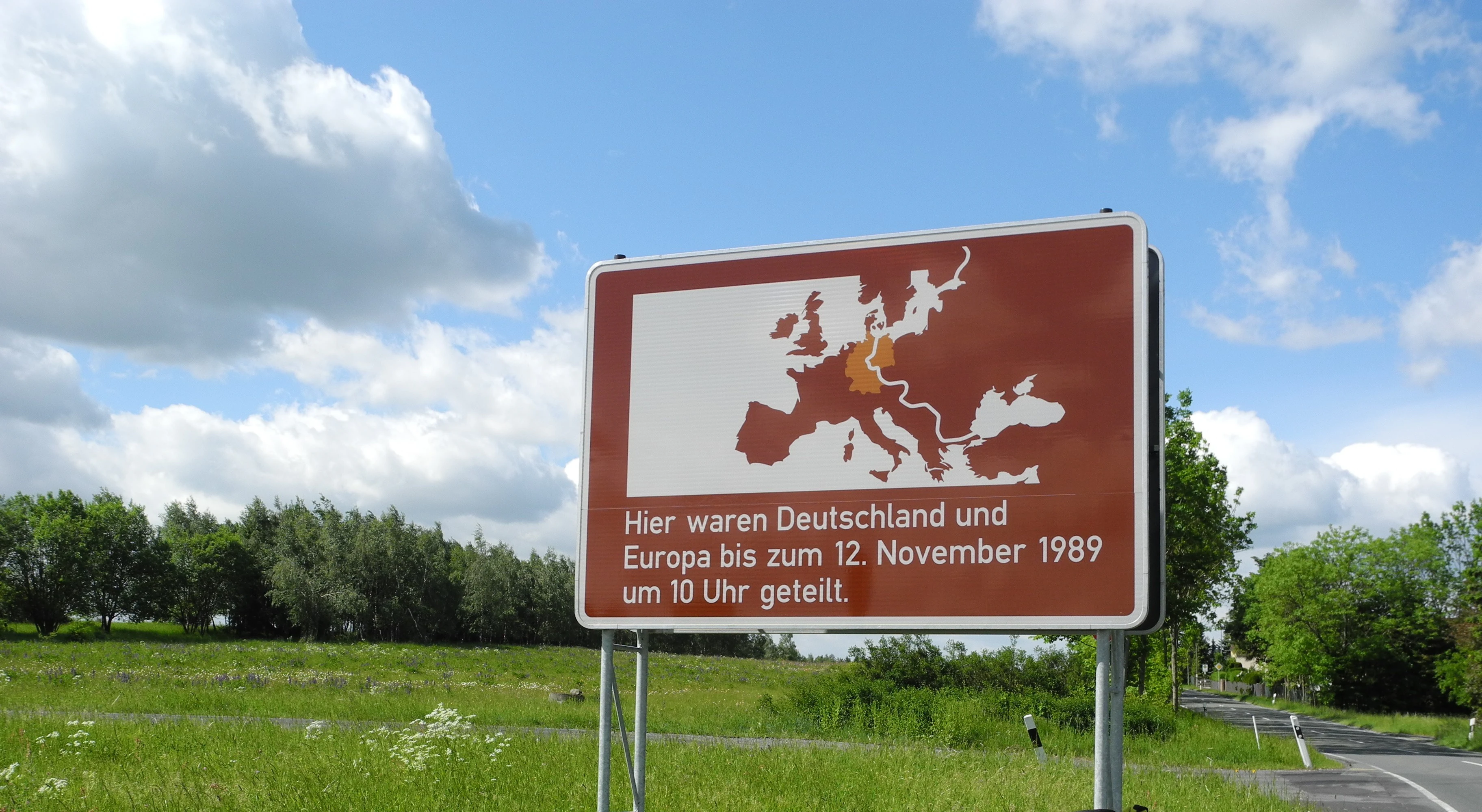 A brown information sign featuring a map of Europe with the former inner-German border highlighted, set against a green field and blue sky with clouds.