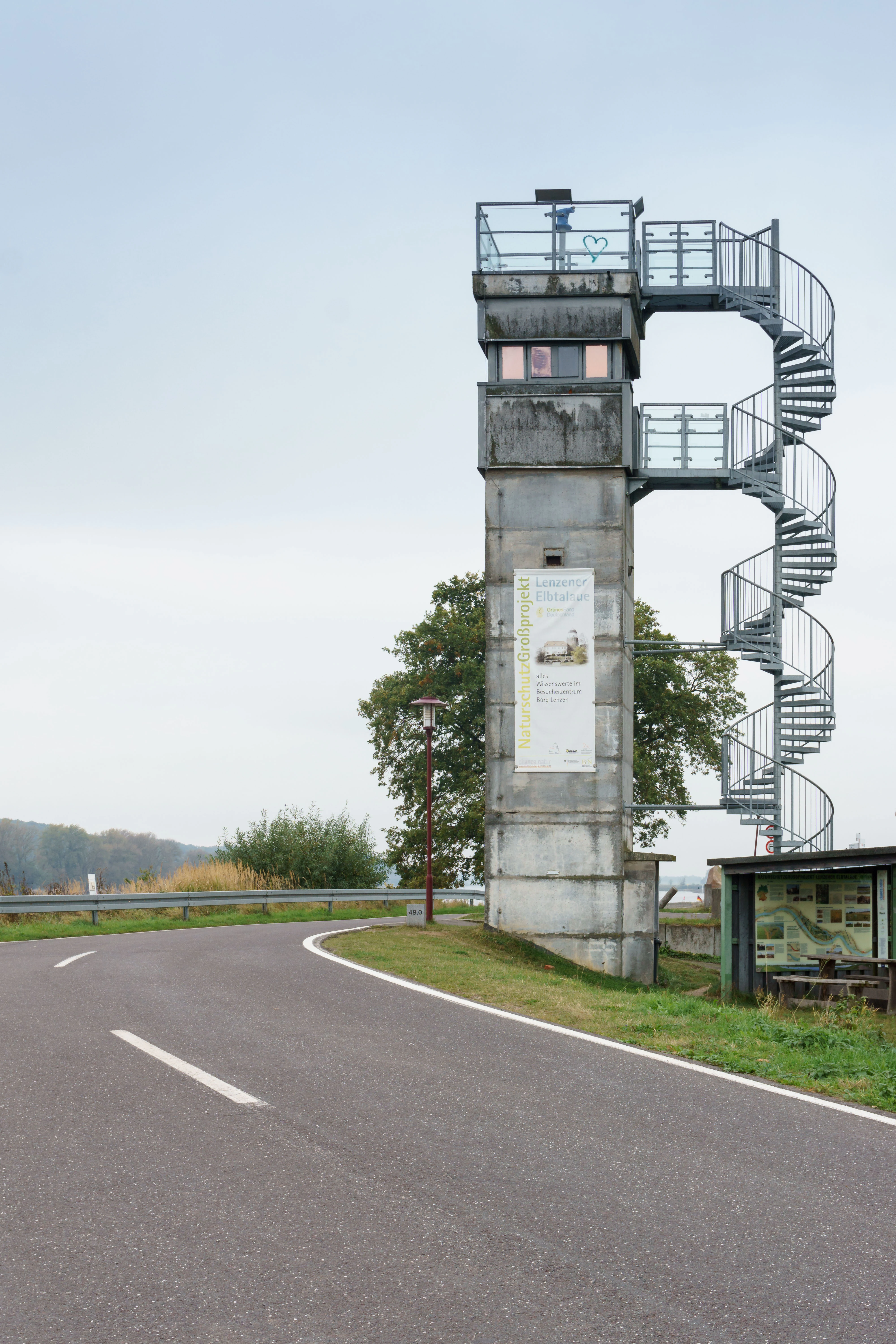 A tall, rectangular concrete watchtower with a metal spiral staircase wrapped around it, located beside a winding asphalt road, with a visitor information banner attached to the side.