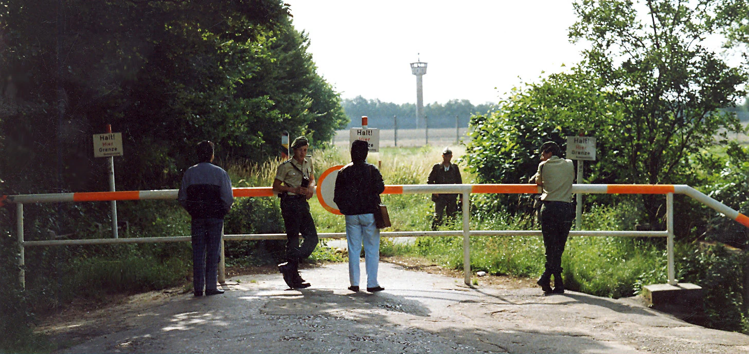 A historical photograph showing a white and orange road barrier blocking a rural path, with several people standing and talking near the gate and a border watchtower visible in the background.