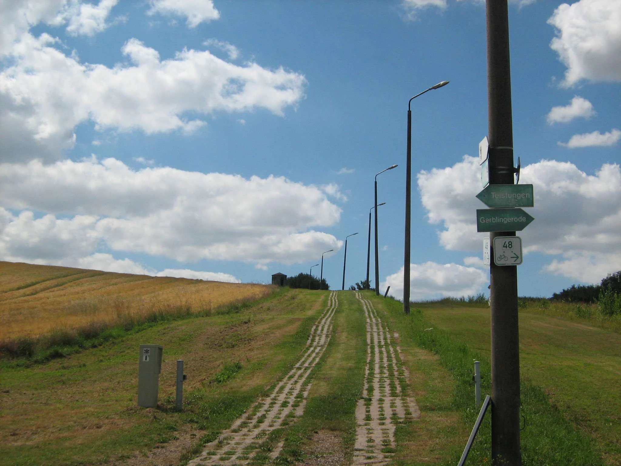 A dirt and concrete path, formerly a border patrol road, stretches uphill through a green, grassy landscape with streetlamps lining the right side.