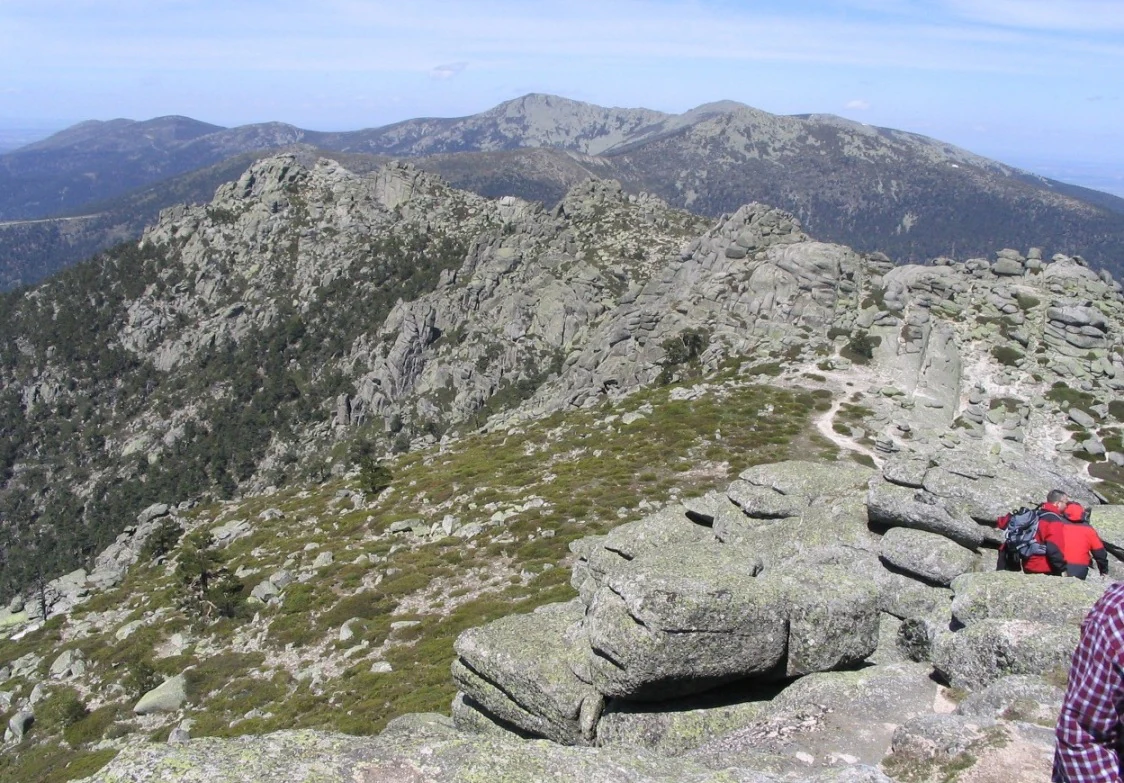 Rocky mountain peaks with green vegetation, a hiker with red backpack on a rocky outcrop, and a winding trail in the background