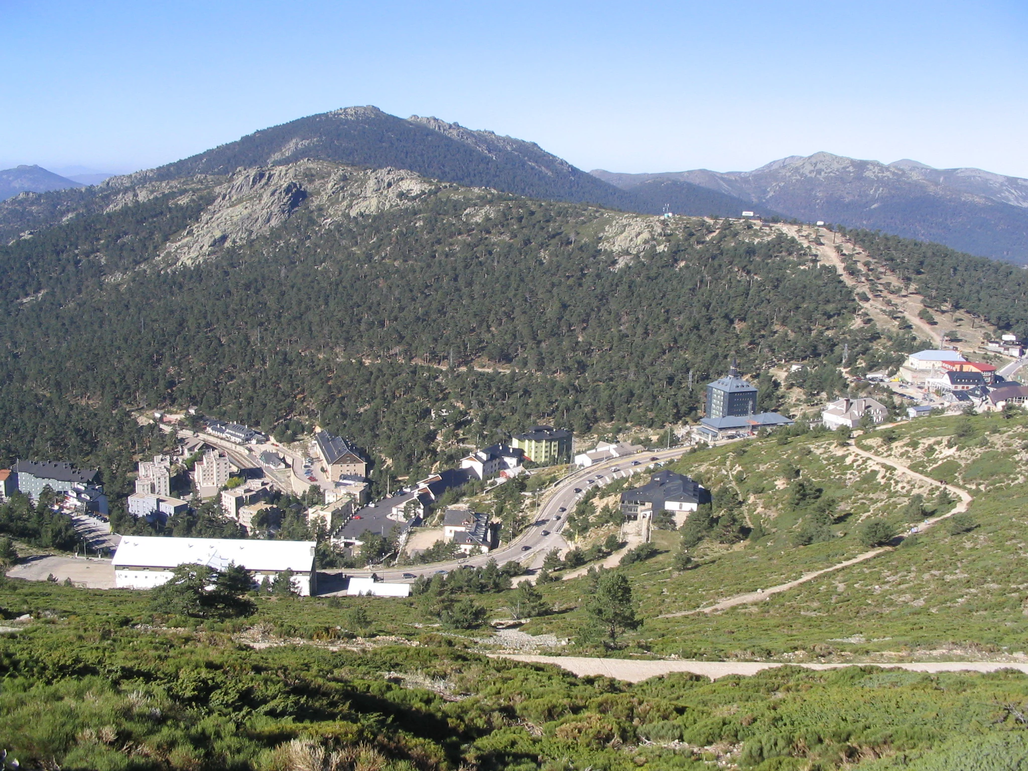 Panoramic view of Puerto de Navacerrada mountain pass showing buildings, roads, and mountainous landscape with green vegetation.