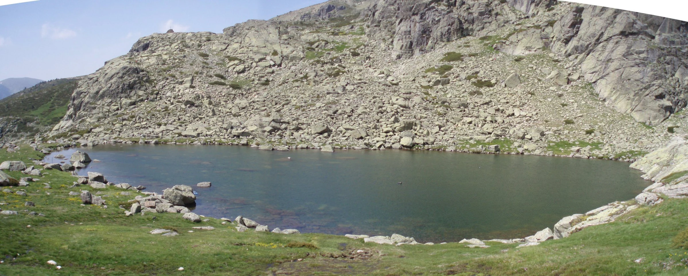 A large alpine lake surrounded by rocky mountains and grassy terrain under a clear sky.