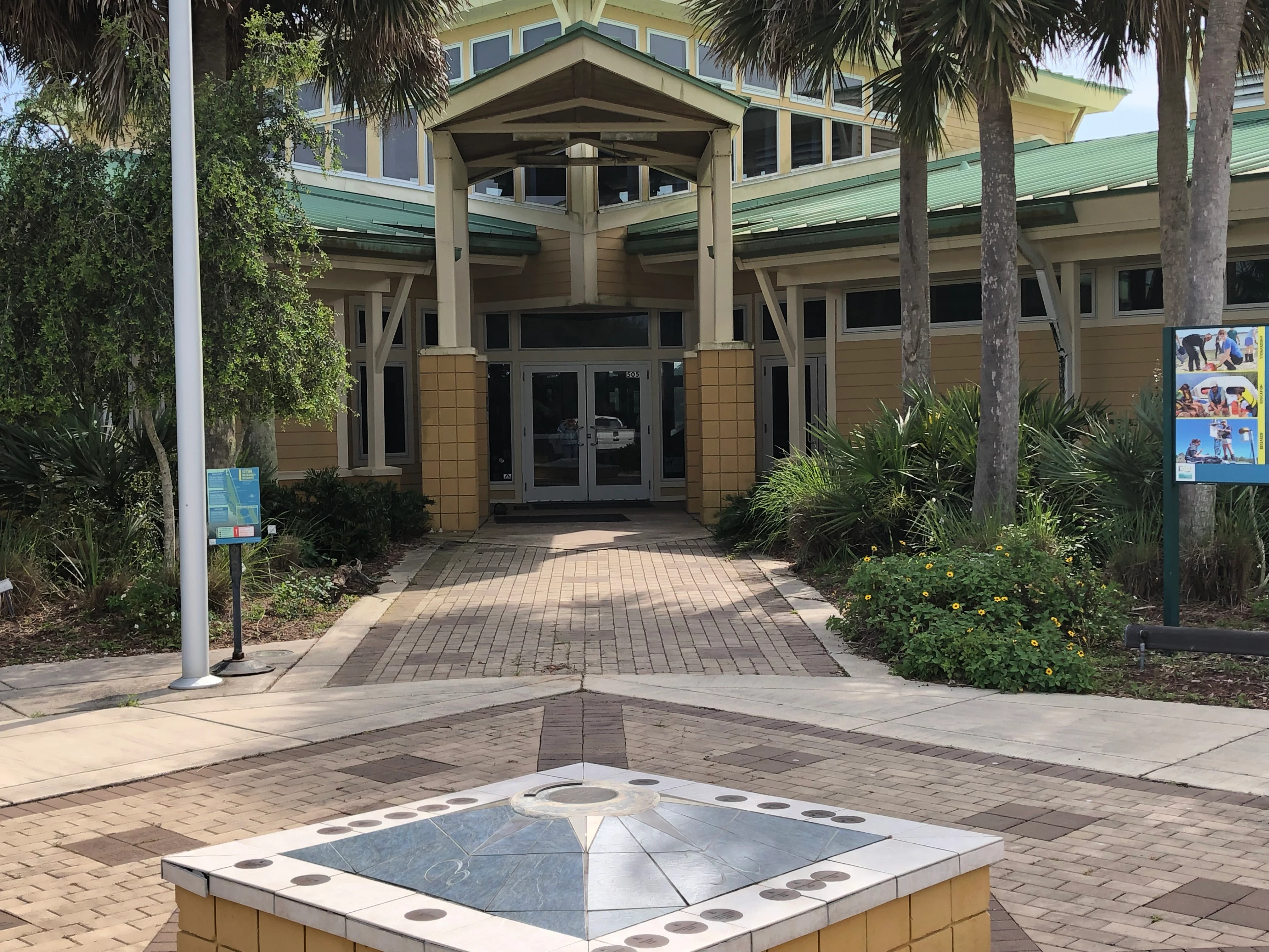 The entrance to the visitor center building at Guana Tolomato Matanzas National Estuarine Research Reserve, featuring a paved walkway, a compass-themed tiled pedestal in the foreground, and palm trees.