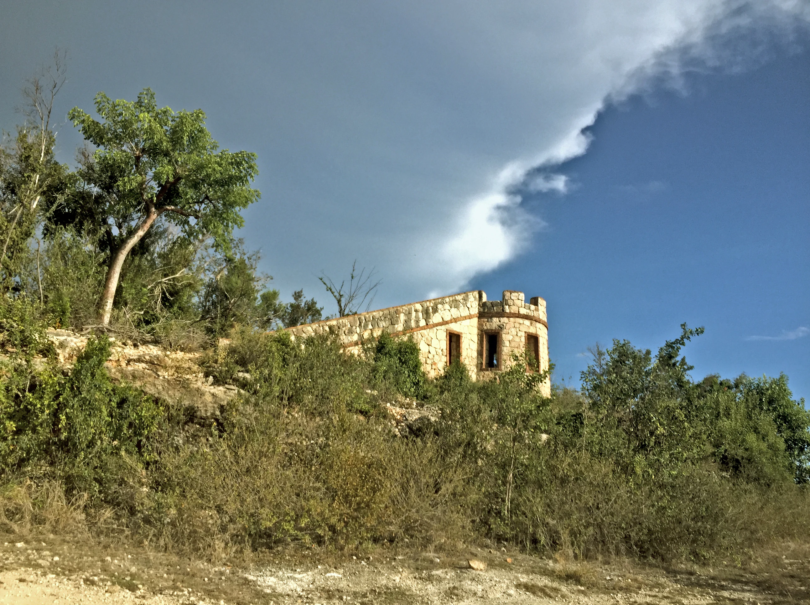 A historic stone fort with crenelated walls stands on a hillside surrounded by dry, scrubby vegetation under a partly cloudy sky.