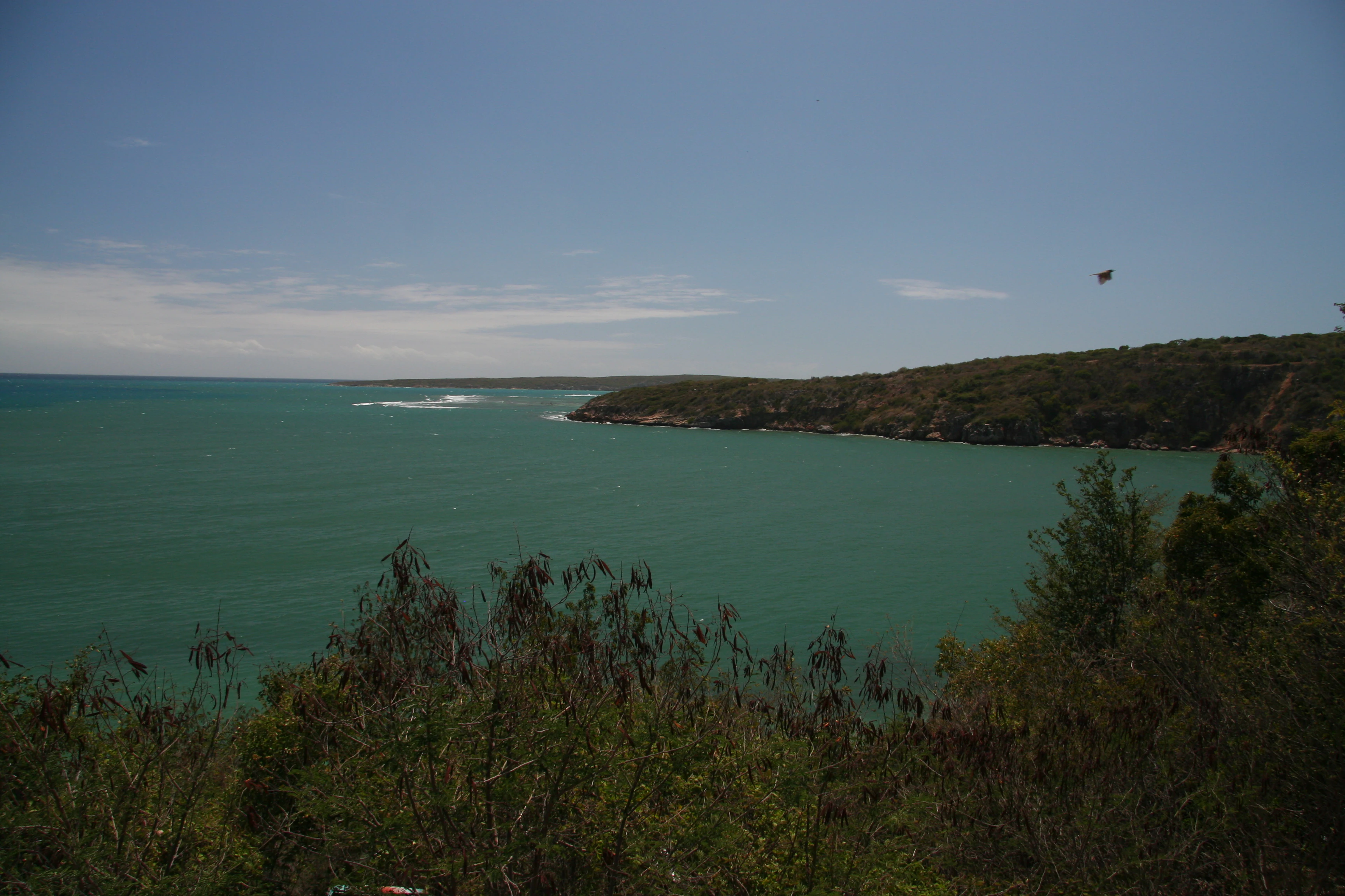 A wide coastal view showing a turquoise bay surrounded by dry, shrubby hills under a clear blue sky.