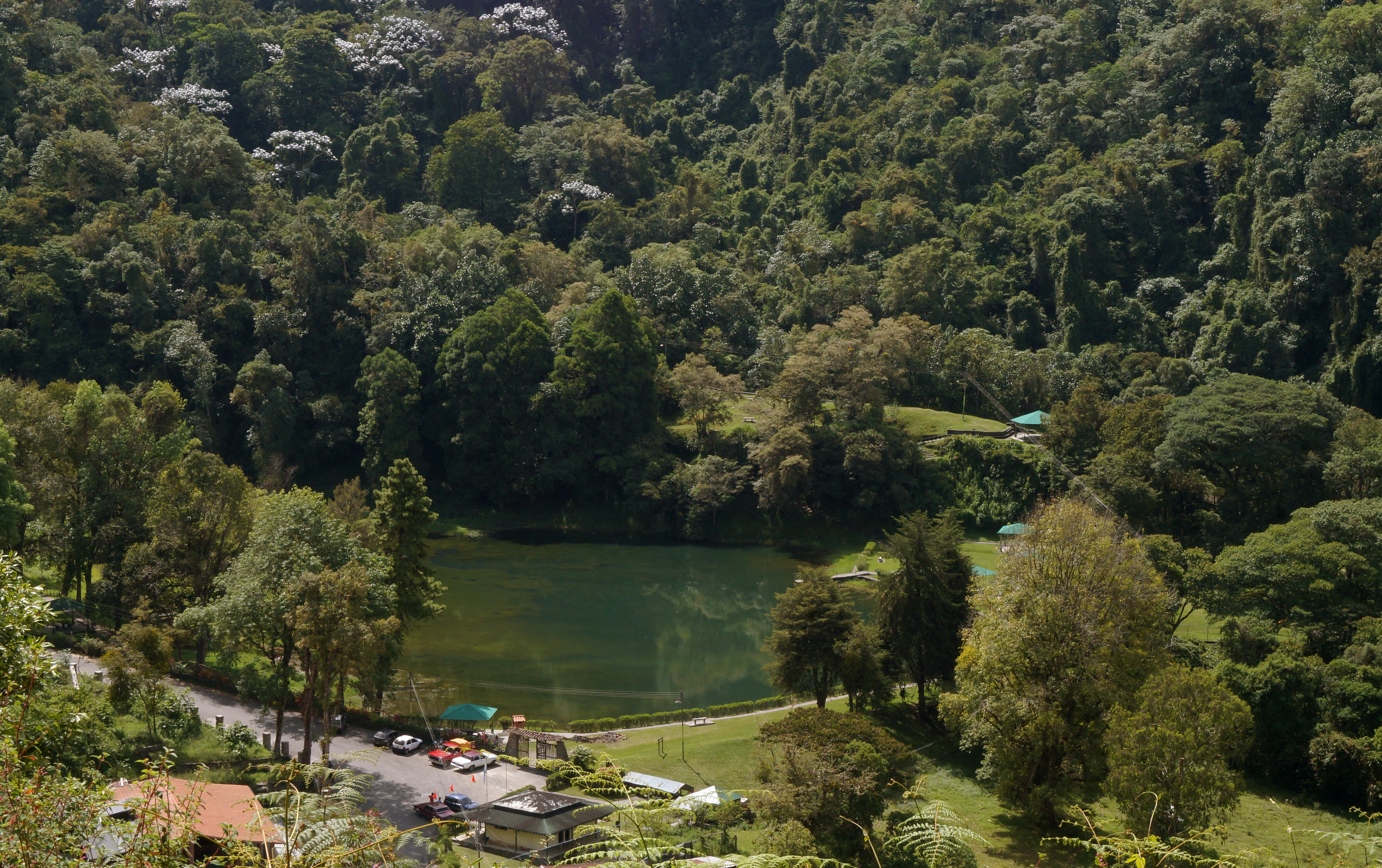 A high-angle view of a calm green lake nestled in a dense, lush mountain forest with a small parking area and structures visible near the shore.