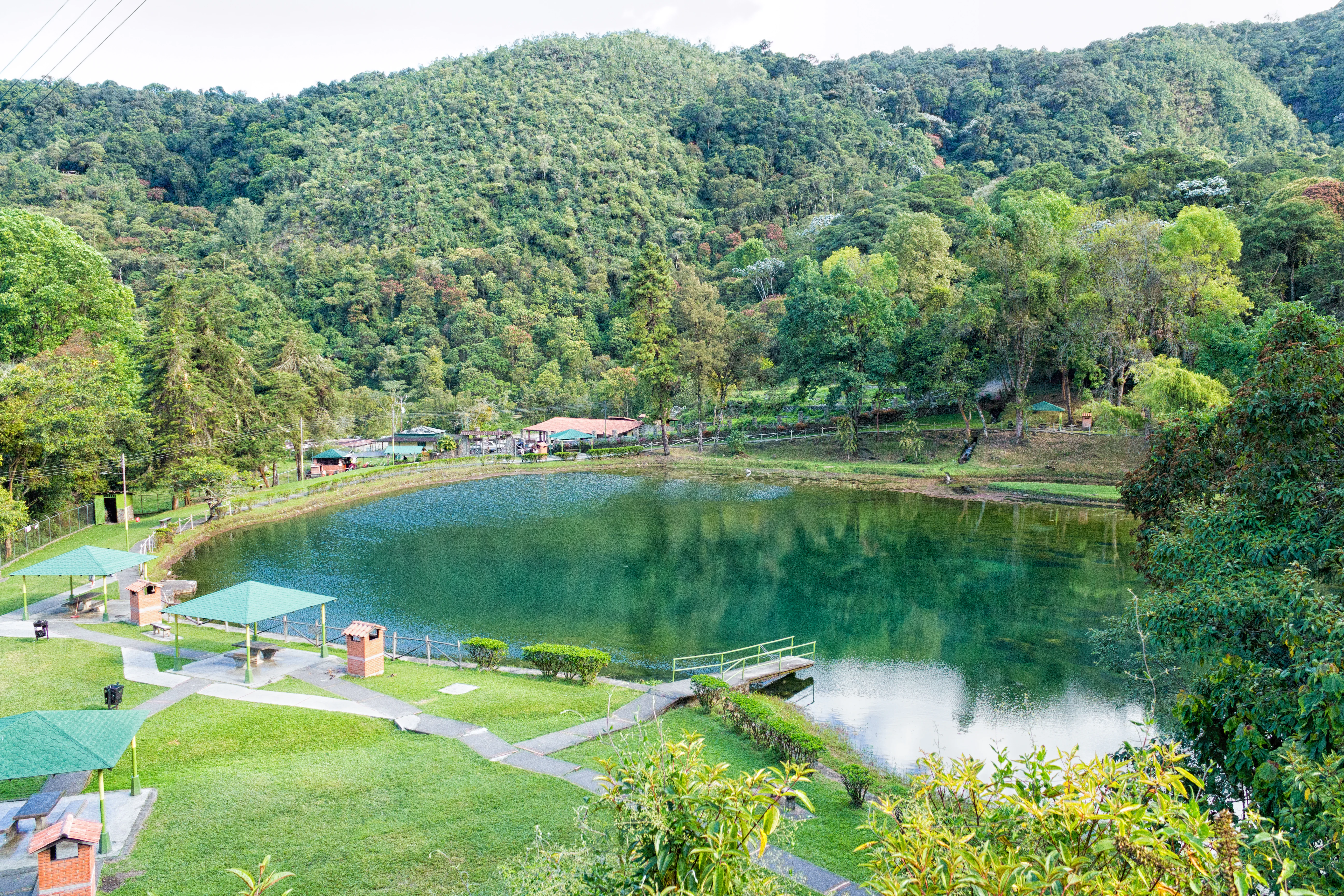 A peaceful green lake reflecting the surrounding dense forest and steep mountain slopes, with picnic shelters and walking paths on the grassy shore in the foreground.
