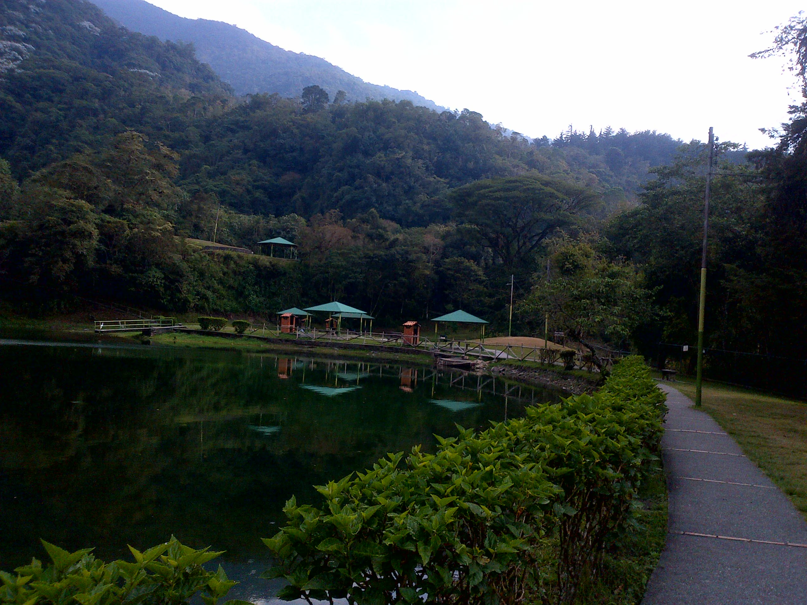 A peaceful lake surrounded by dense, forested mountains with a paved walkway and green-roofed picnic shelters along the shore.