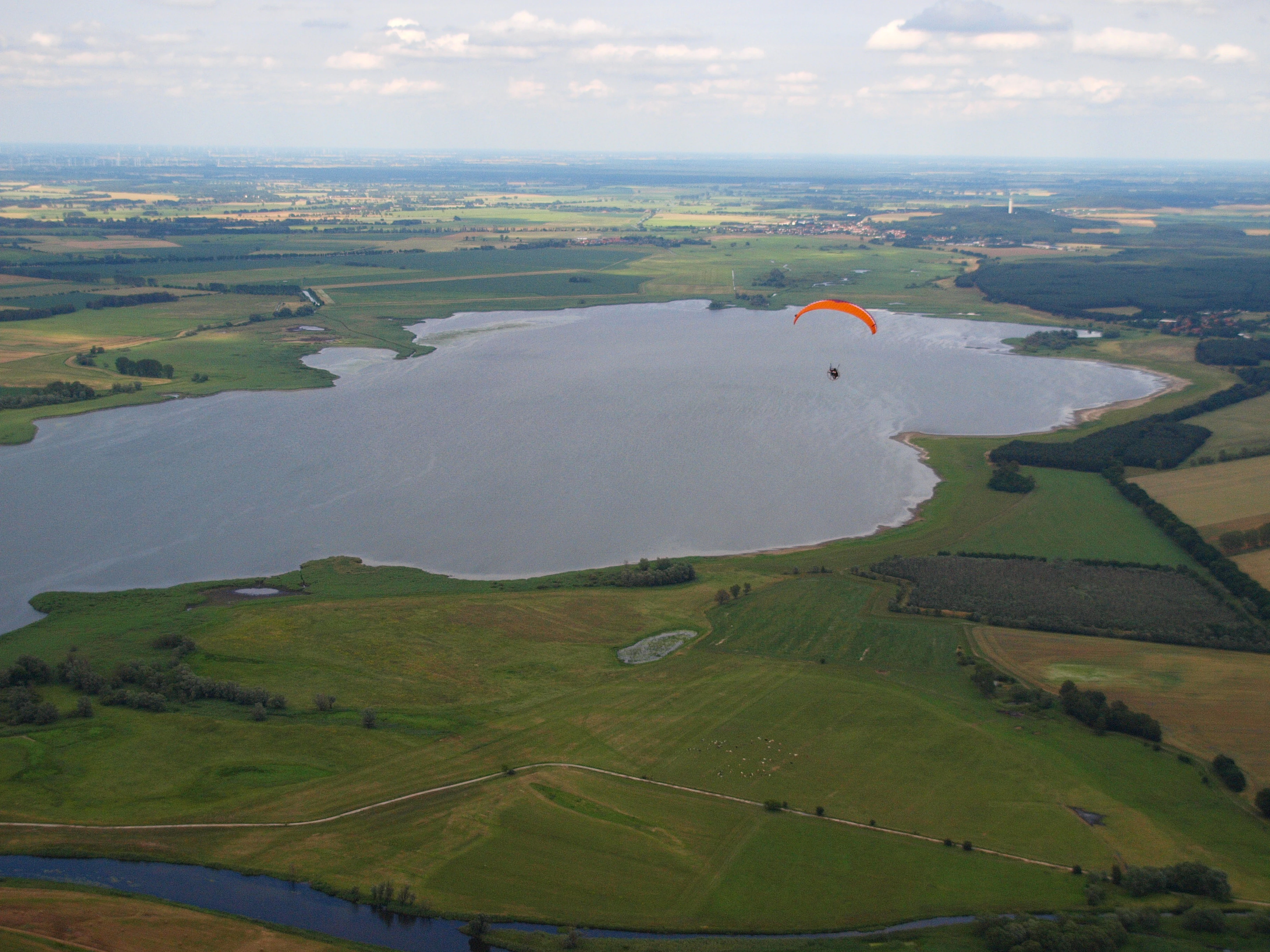 Una vista aérea del lago Gülper See en Brandeburgo, rodeado de campos agrícolas y paisaje verde, con un parapente visible sobre el agua.