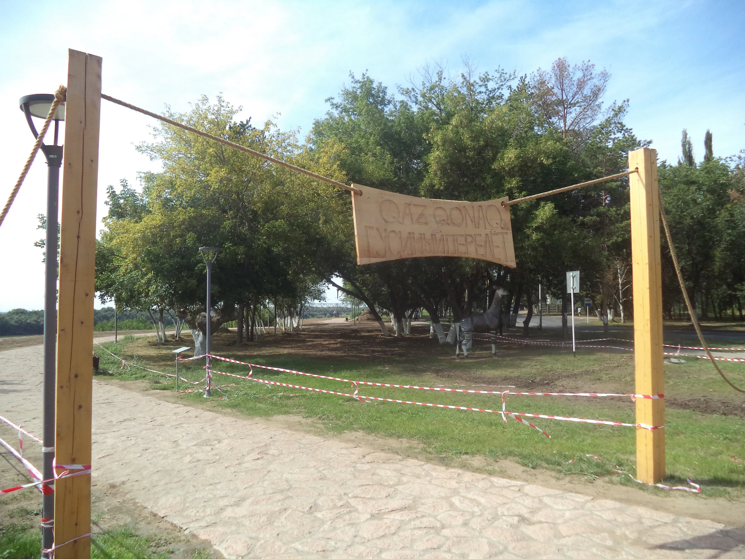 A wooden entrance gate with two tall posts and a hanging banner, with red and white caution tape blocking a dirt path leading into a wooded area.