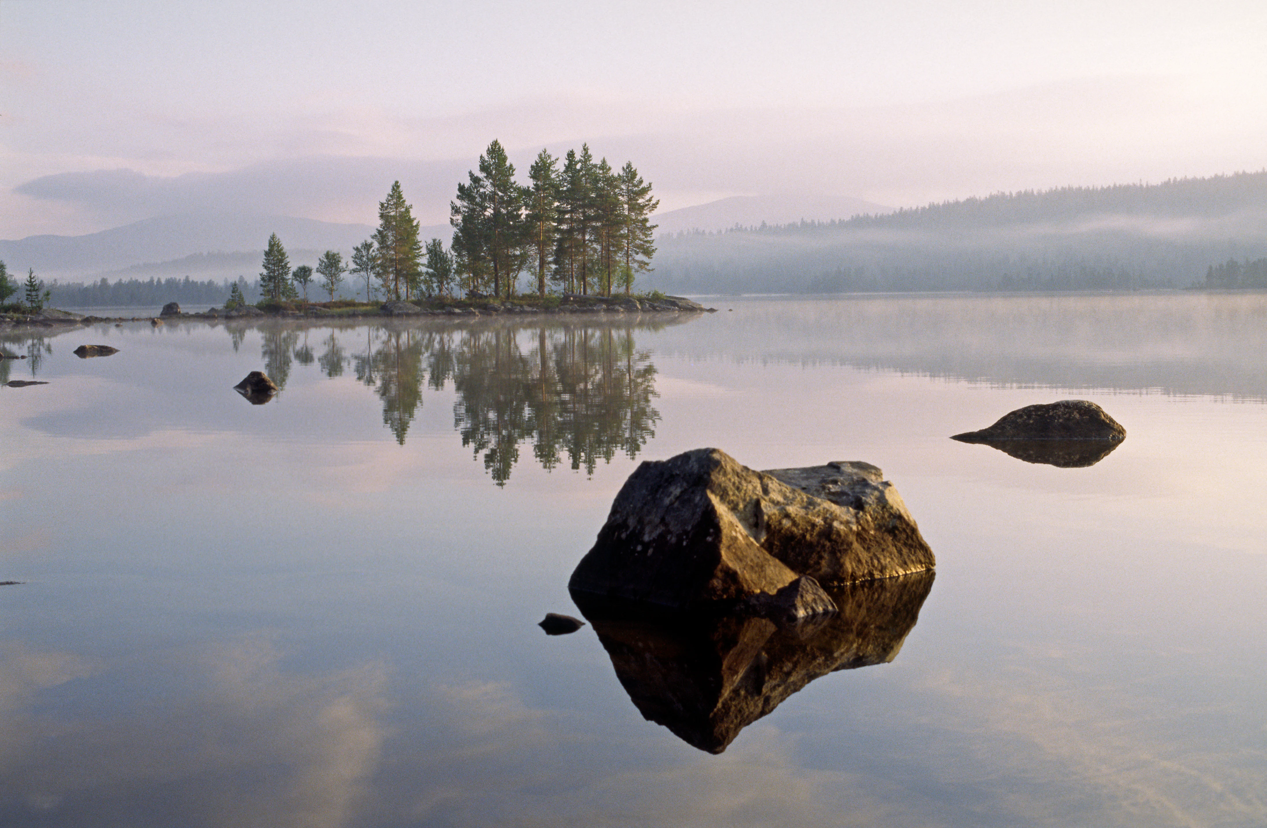 Calm lake reflecting misty mountains and trees with rocks in the foreground
