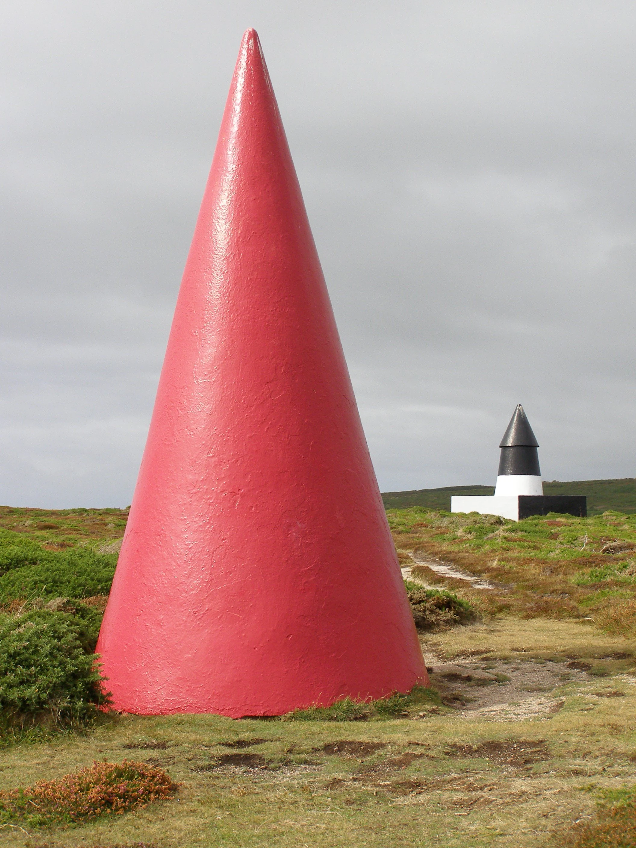 A large, bright red conical navigation daymark stands in the foreground, with a smaller black and white conical daymark visible in the grassy distance under an overcast sky.