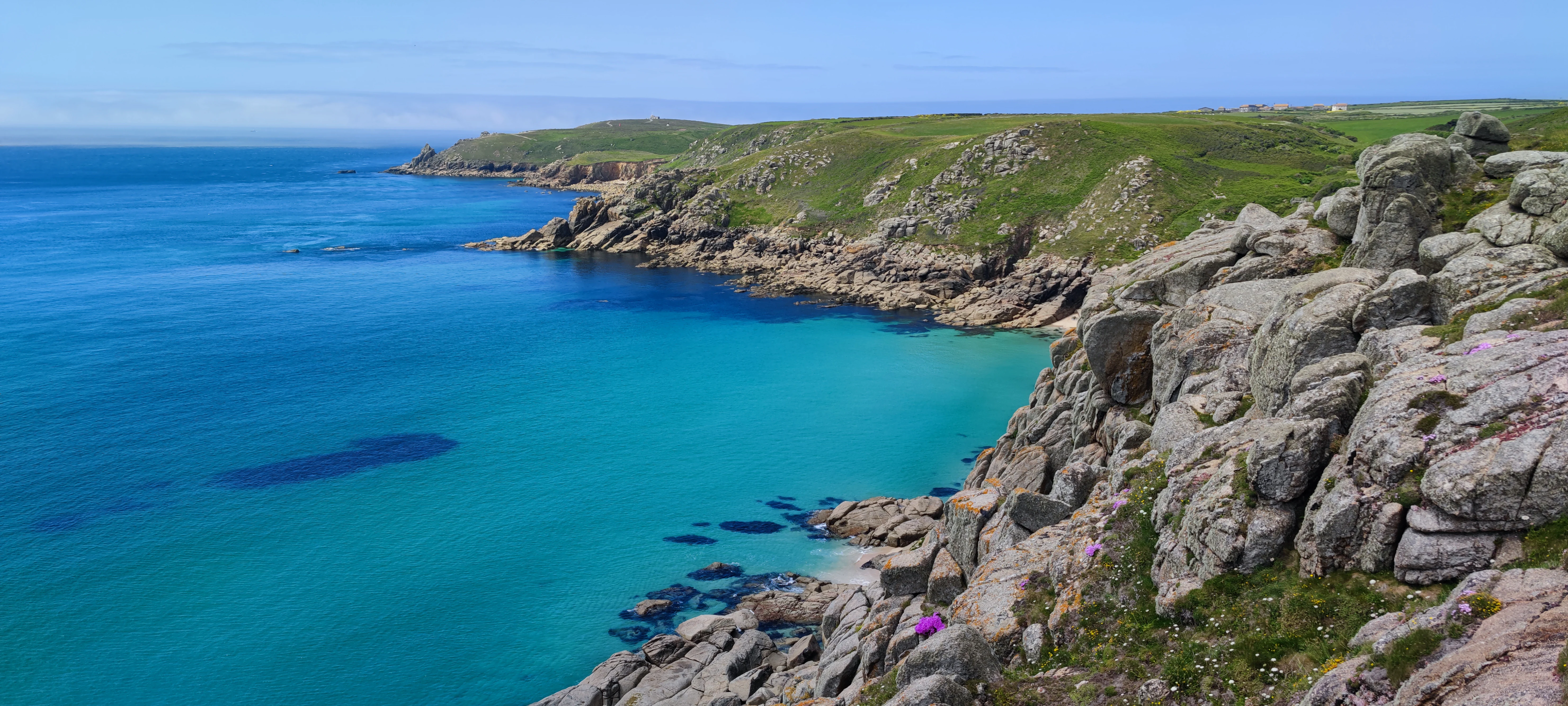 A high-angle view of rugged granite cliffs overlooking a turquoise sandy cove, with grassy headlands stretching into the distance along a coastline.
