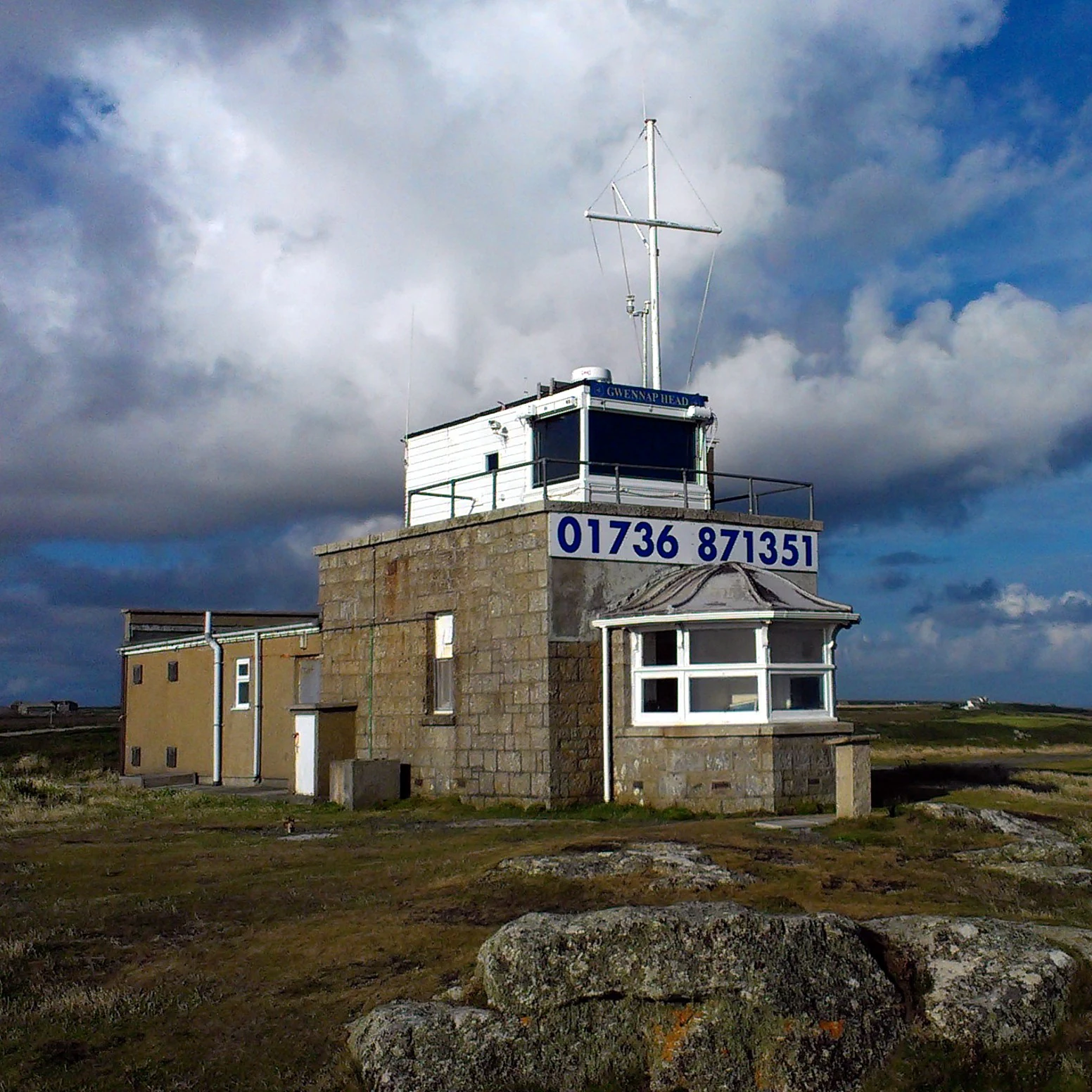 A small, stone-built lookout building with a white upper cabin and communications mast on a rocky, grassy headland under a cloudy sky.