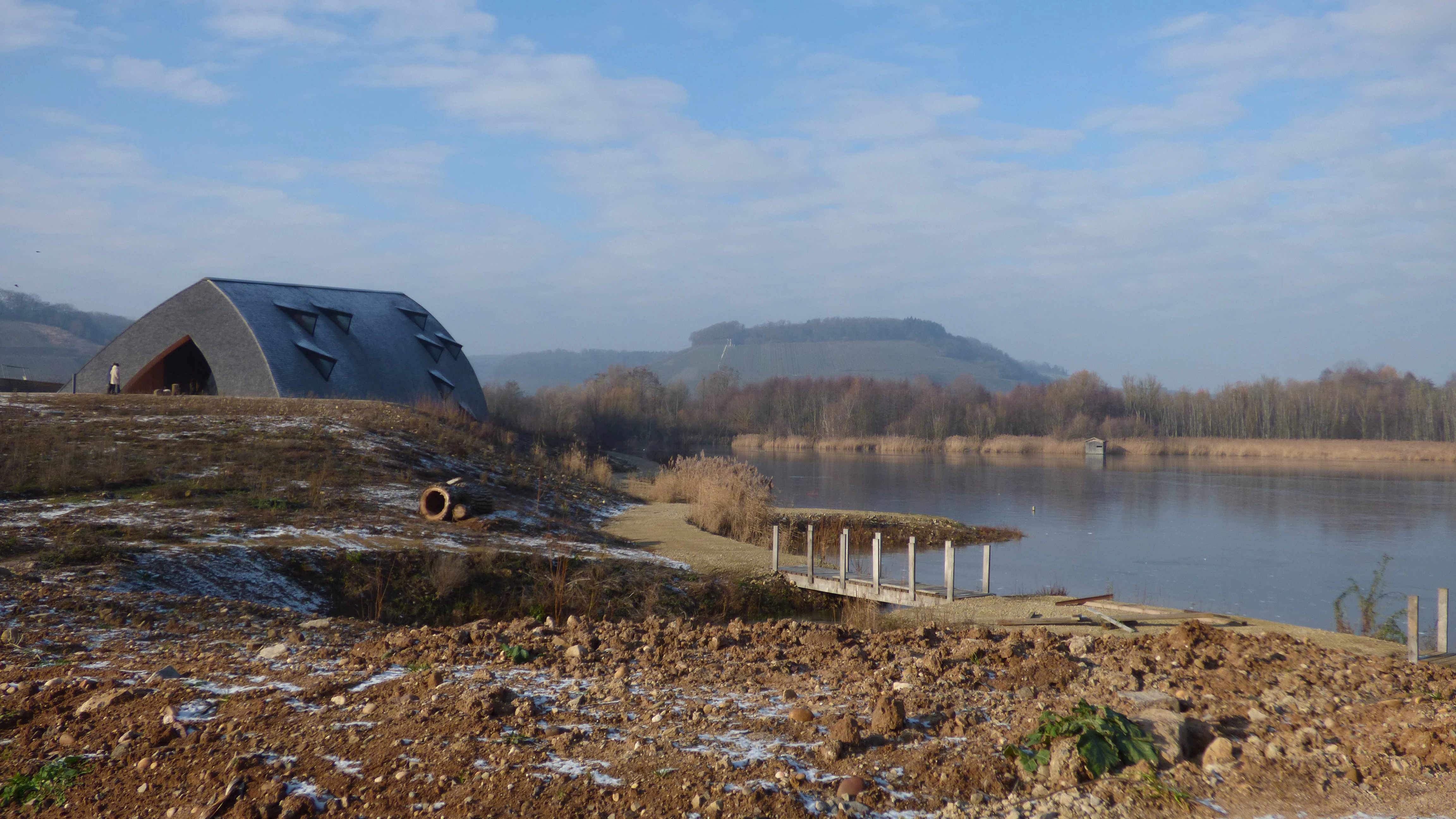 A modern curved structure stands on a grassy bank next to a calm lake, with hills in the background on a bright winter day.