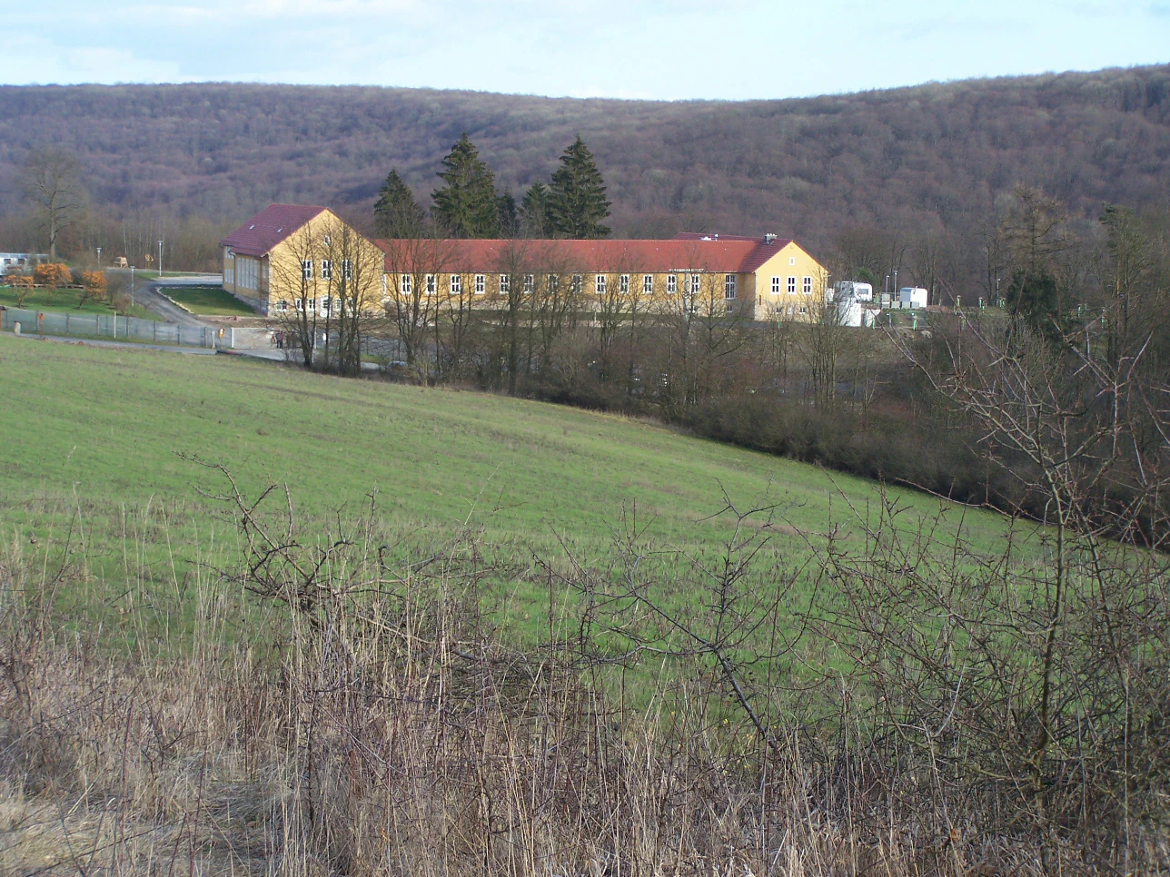 A long, yellow, one-story building with a red roof sits in a grassy clearing with a forested hillside in the background.