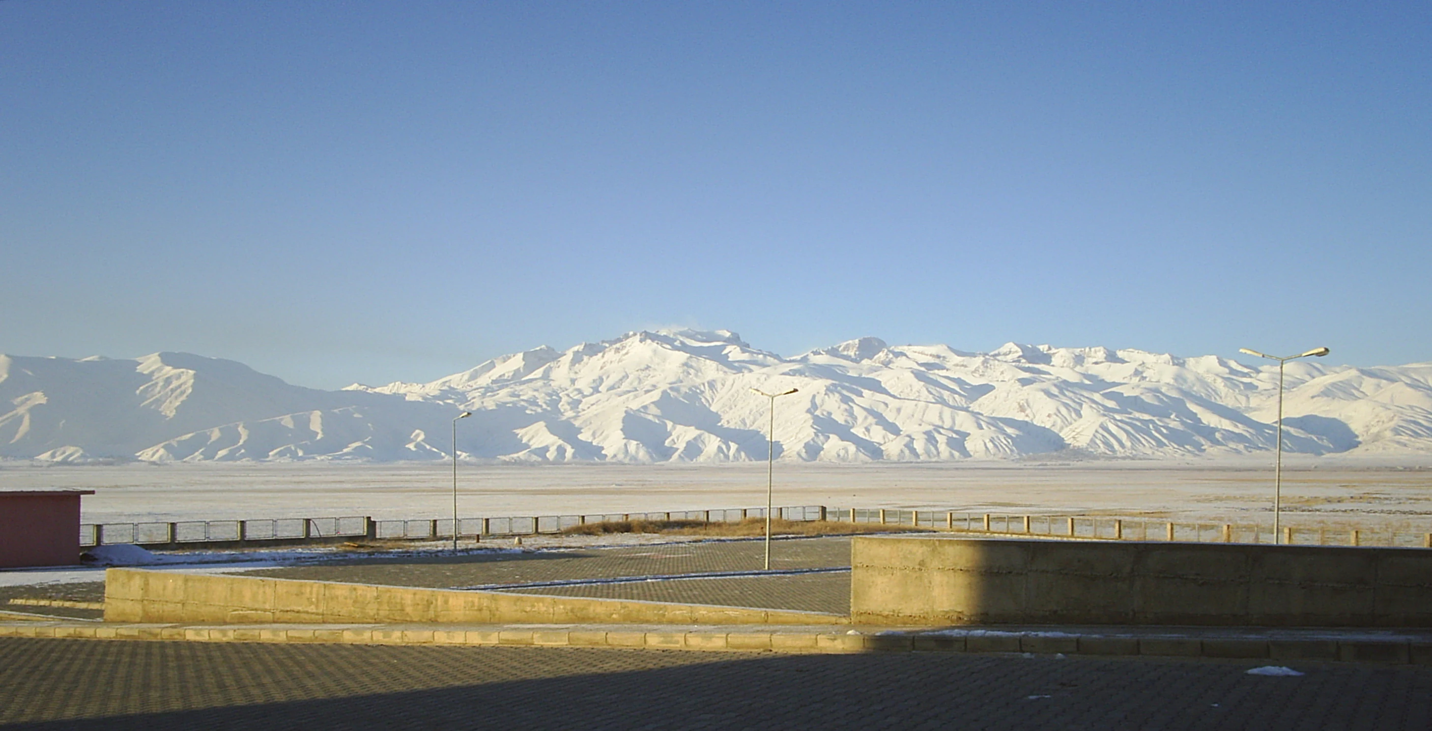 Wide landscape view of snow-covered mountain peaks under clear blue sky with a concrete structure and paved area in the foreground
