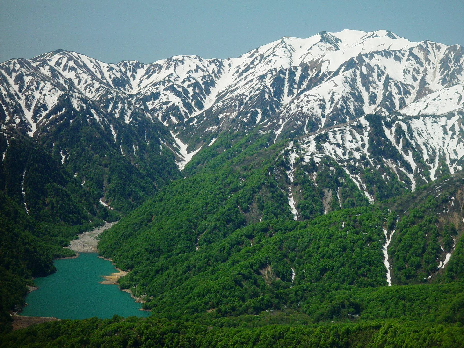 Una vista panorámica de un lago turquesa rodeado de densos bosques verdes y picos montañosos nevados bajo un cielo azul claro