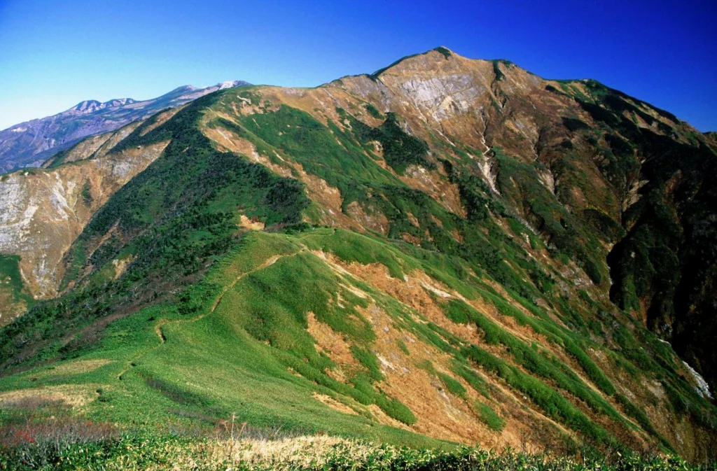 Pendientes montañosas verdes y marrones con afloramientos rocosos bajo un cielo azul despejado.
