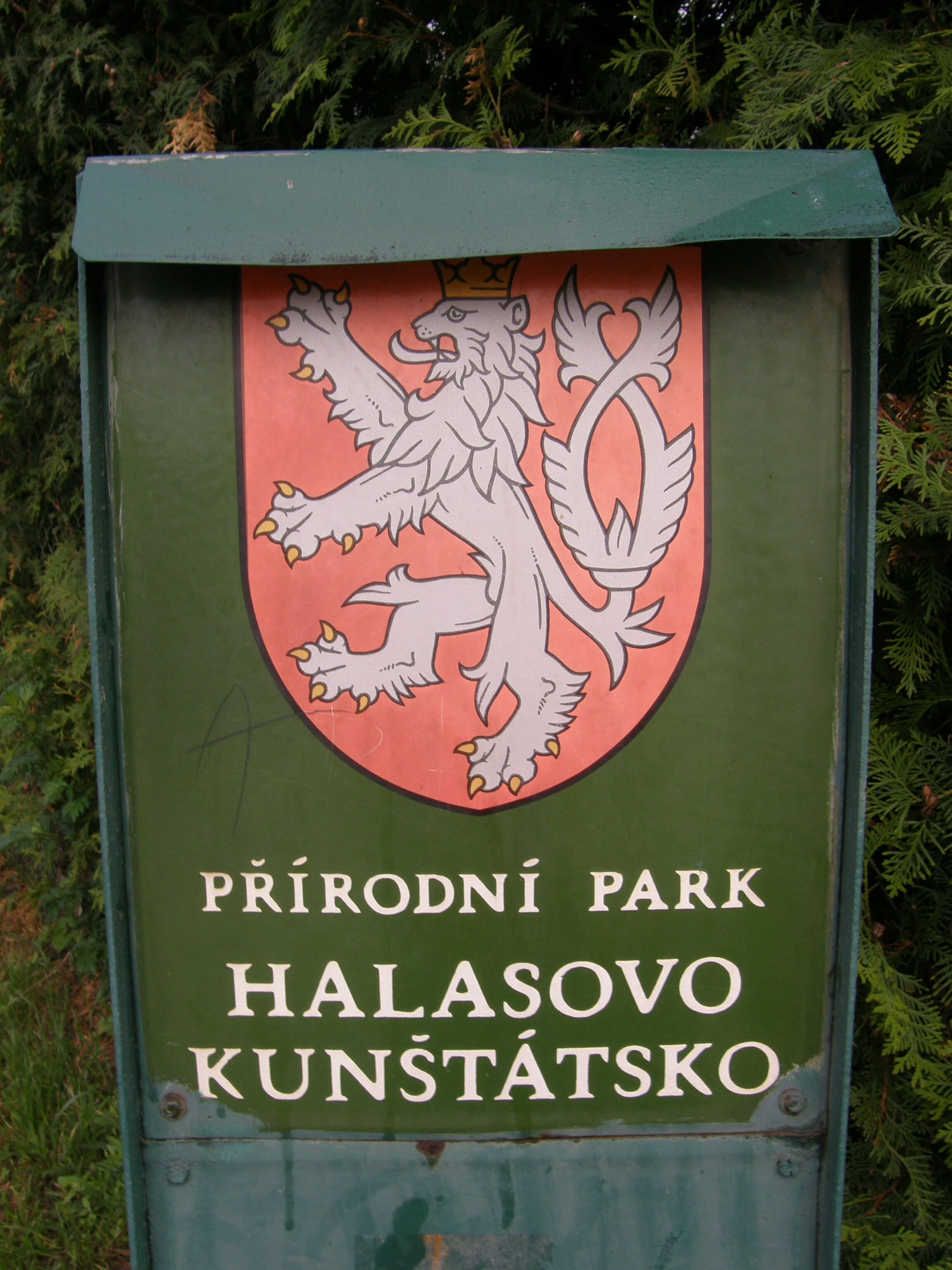 A green metal park information sign displaying the name PŘÍRODNÍ PARK HALASOVO KUNŠTÁTSKO and the Czech heraldic lion emblem.