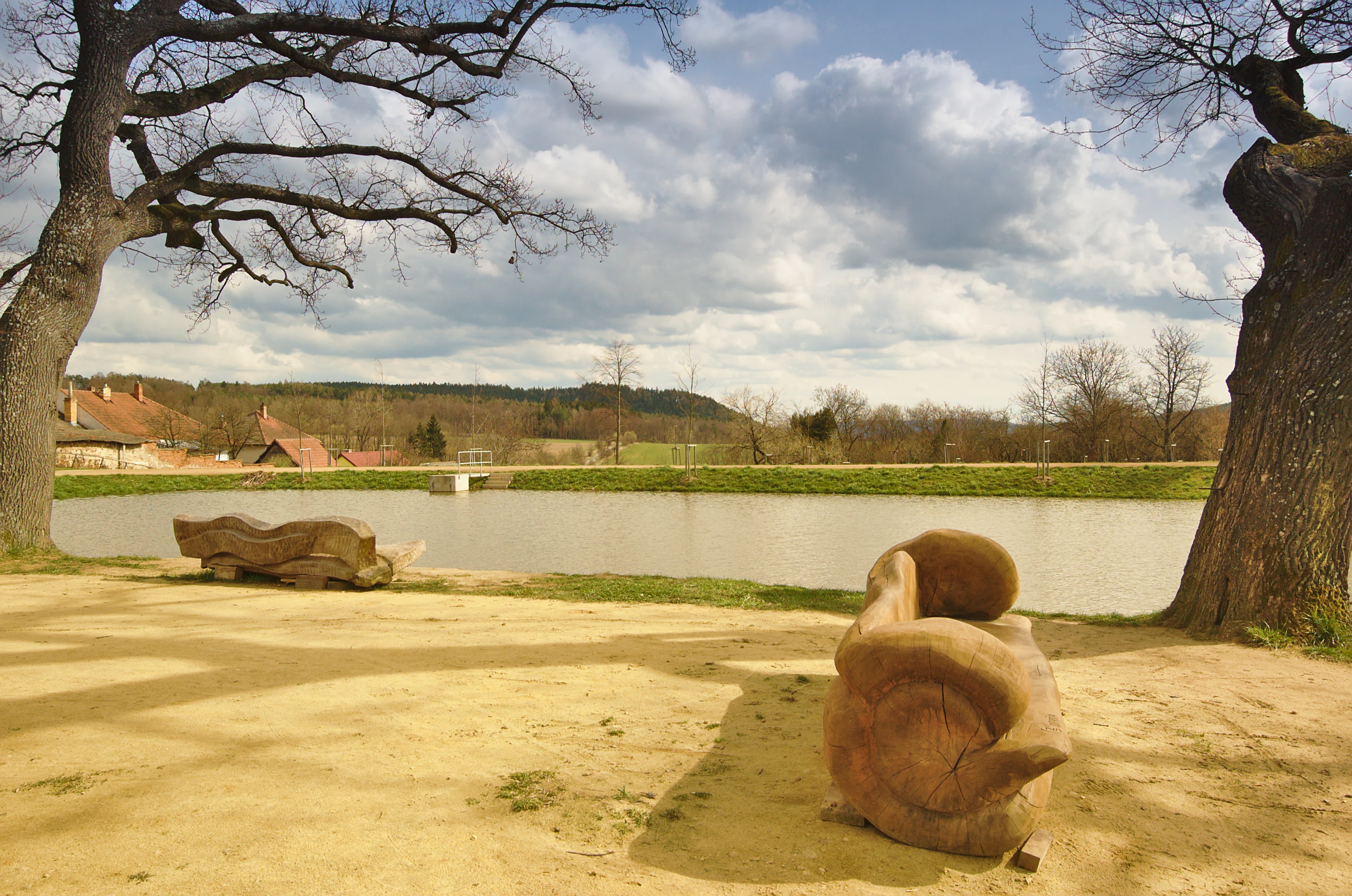 A calm pond surrounded by trees, with large, carved wooden benches resting on a dirt path in the foreground.