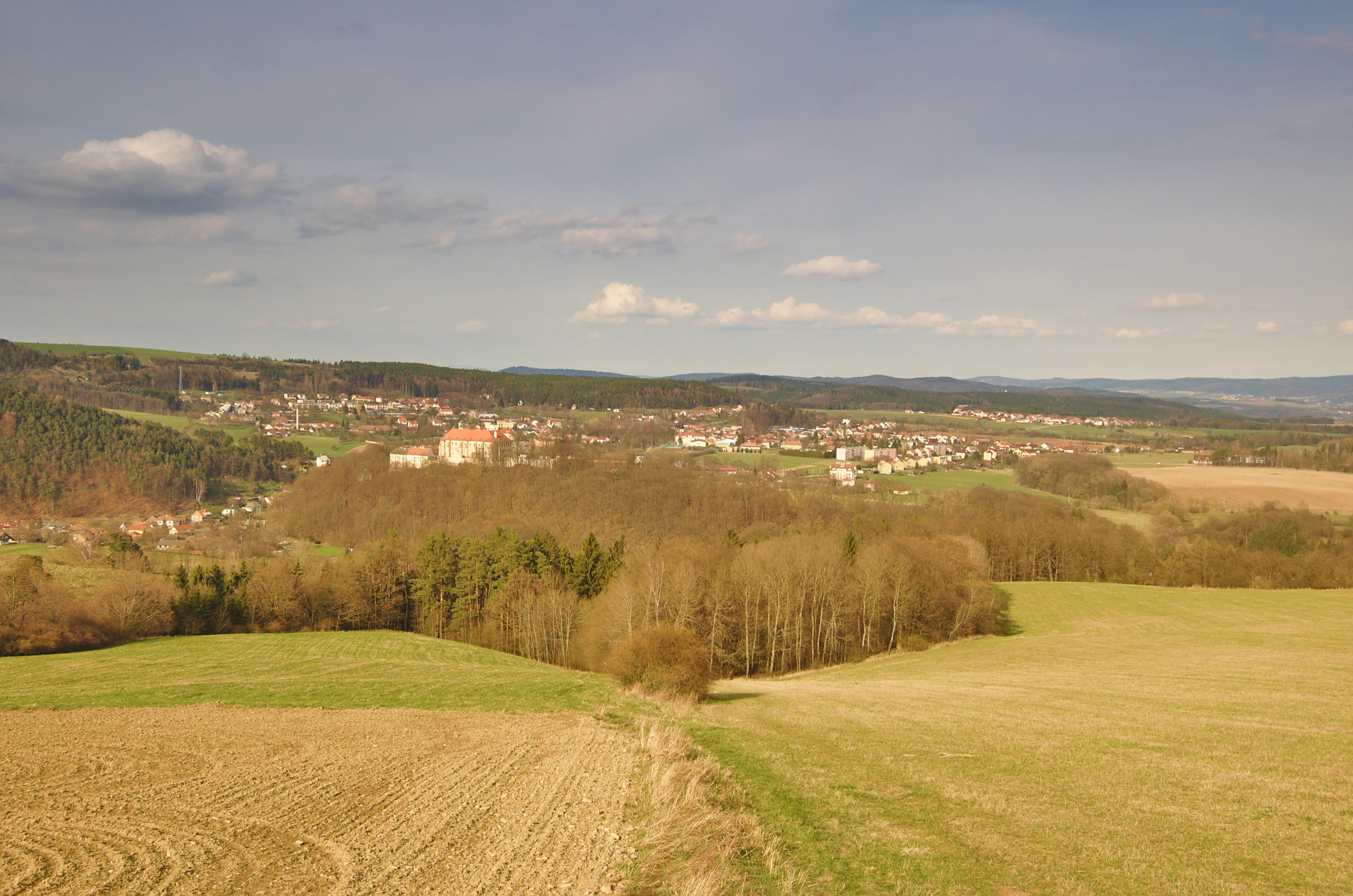 A wide-angle, elevated view of a small town nestled in a valley with rolling hills, meadows, and patches of forest under a light blue sky.