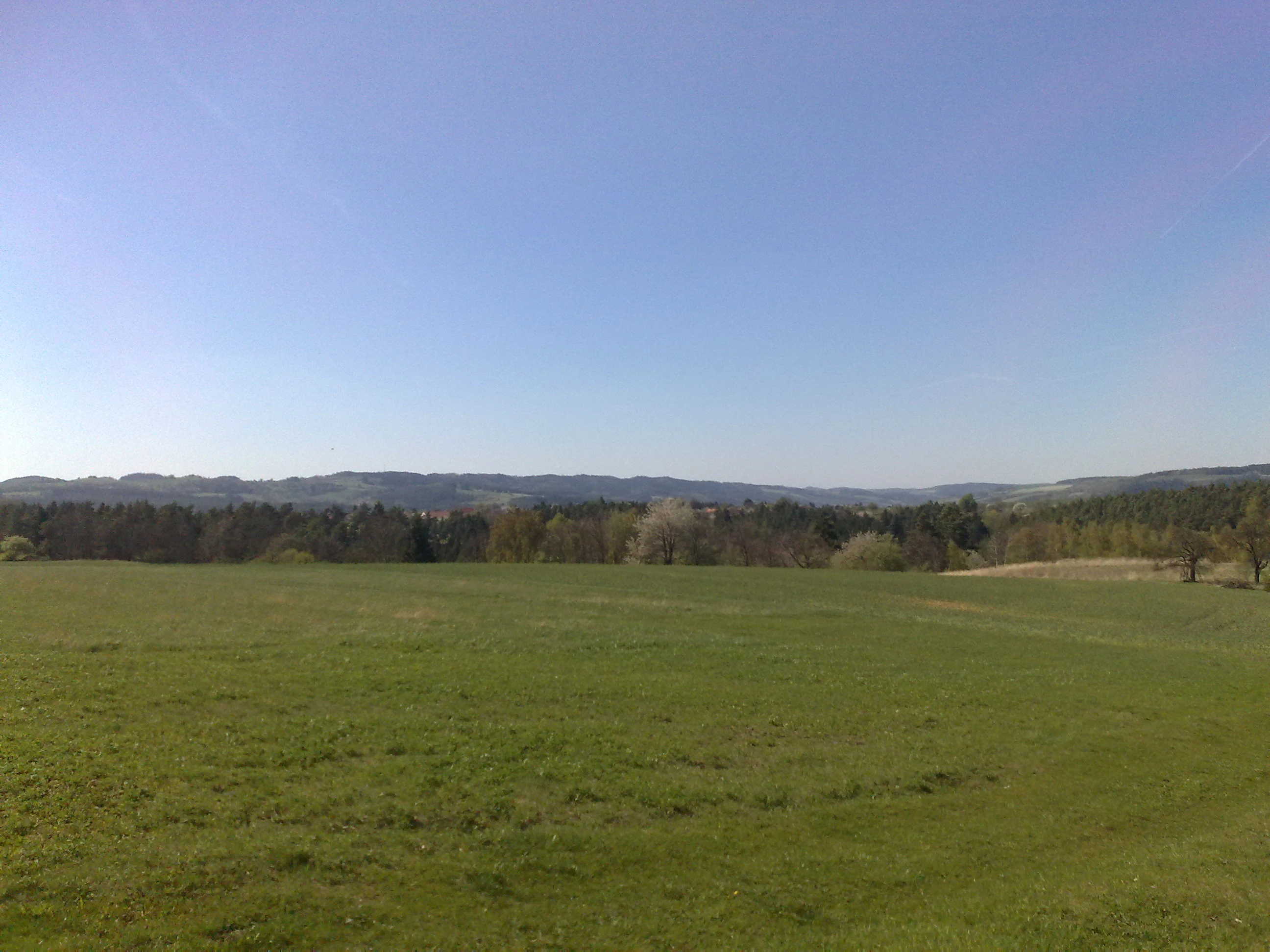 A wide, eye-level landscape shot of a green grassy field in the foreground, with a line of trees and rolling hills in the background under a clear blue sky.