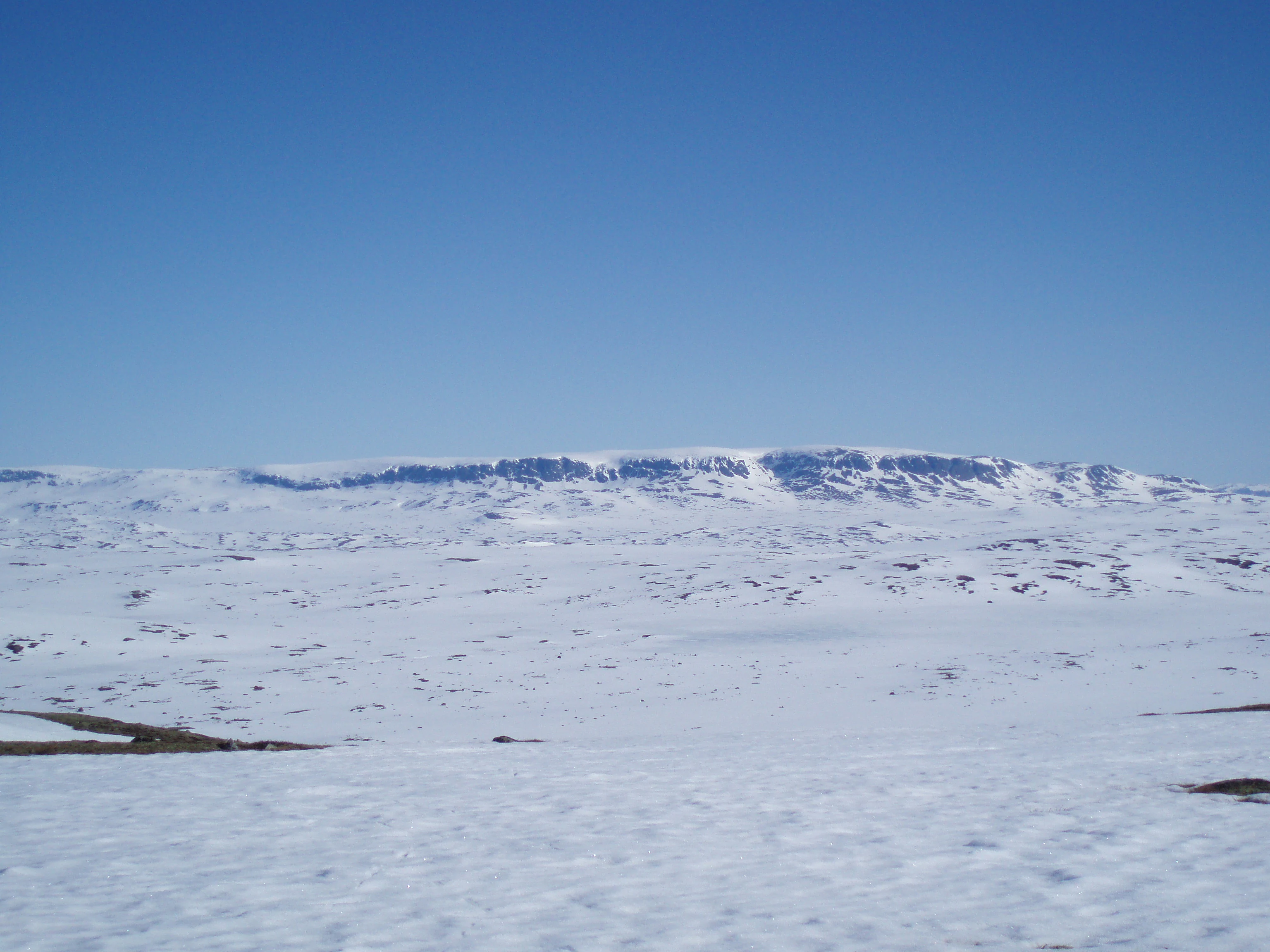 Snow-covered mountain plateau under clear blue sky with distant mountain range