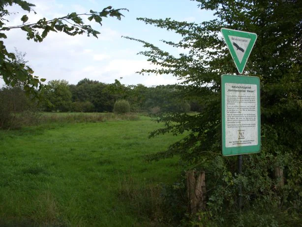 A green-and-white nature reserve sign on a metal post at the edge of a green meadow, with trees in the background.