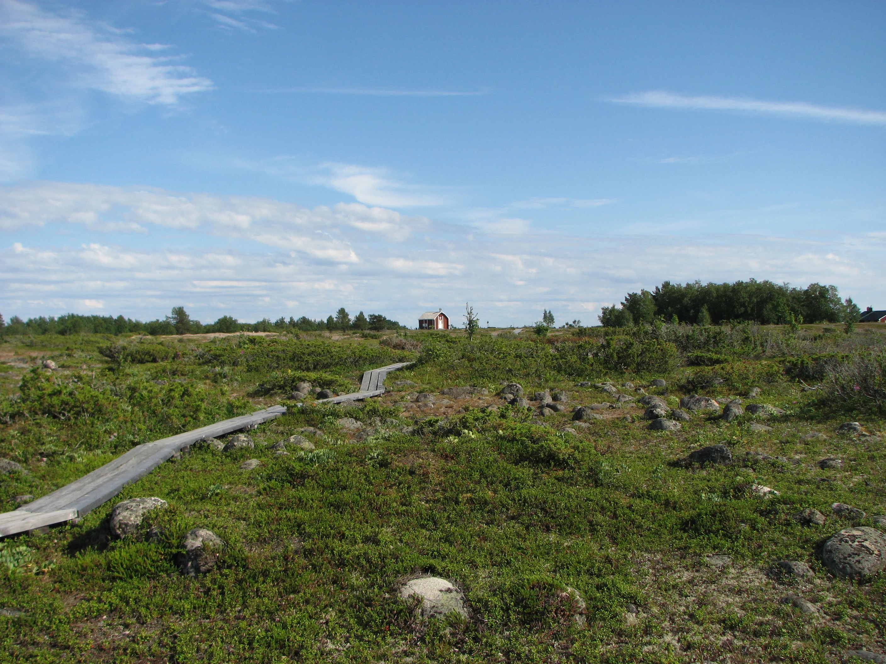 Wide landscape view of Sandskär island showing a wooden boardwalk through rocky terrain with green vegetation, a small red building in the distance, and a clear blue sky
