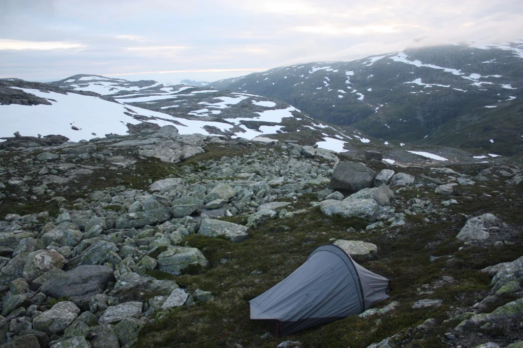 Gray hiking tent on rocky ground with patches of snow and distant mountain peaks under an overcast sky