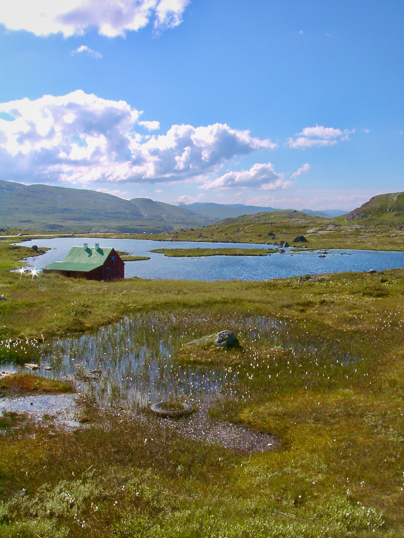 Landscape of Hardangervidda National Park featuring a small red cabin with green roof, a lake, grassy terrain with scattered water pools, and distant mountains under a blue sky with white clouds