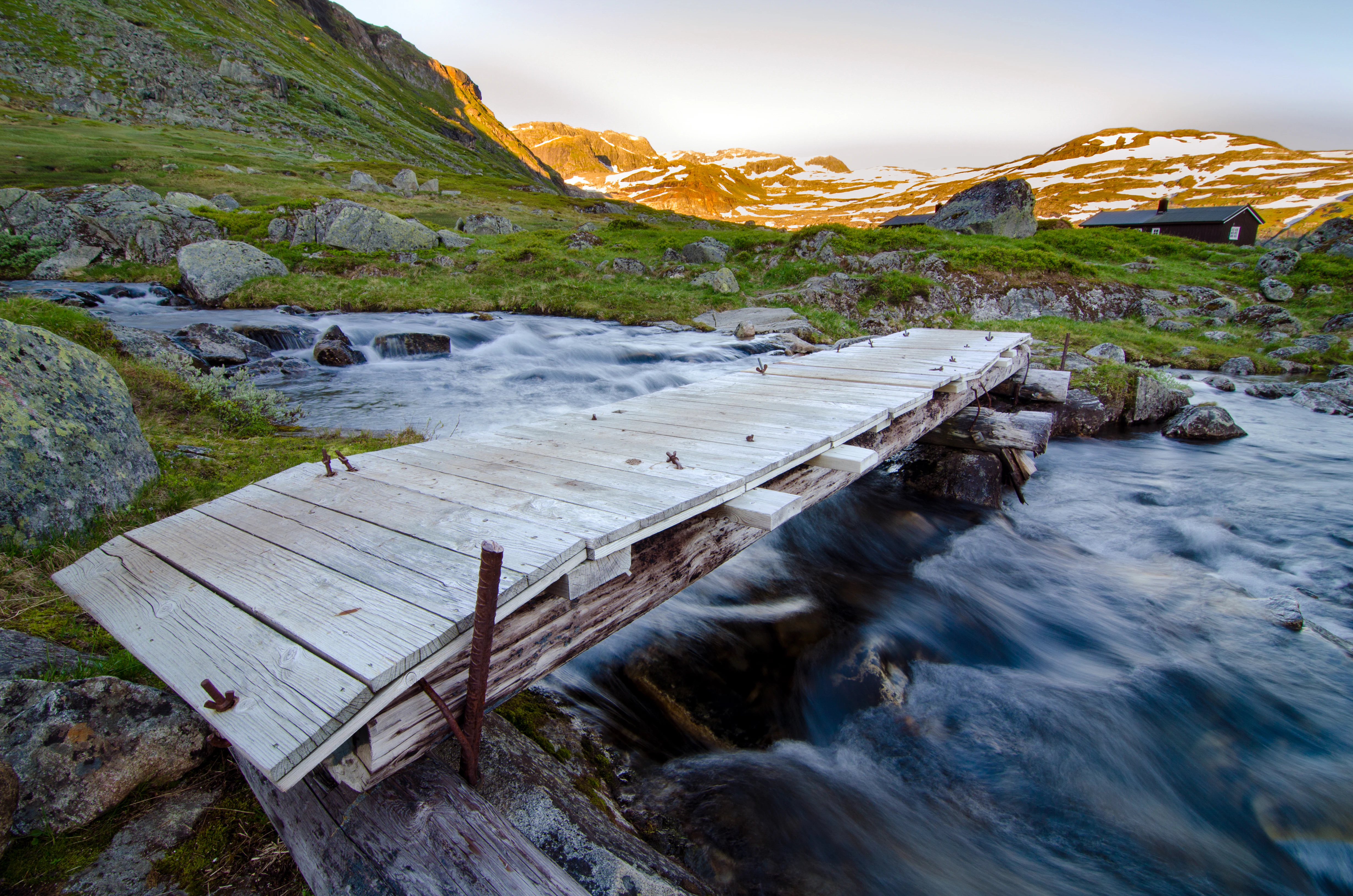 Wooden bridge spanning a flowing river with mountainous landscape under a sunset sky