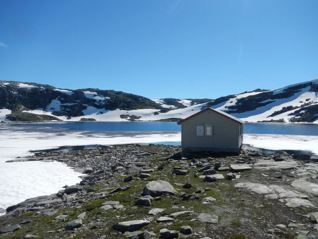 A small gray cabin on rocky terrain with snow patches, a lake, and snow-capped mountains under a clear blue sky