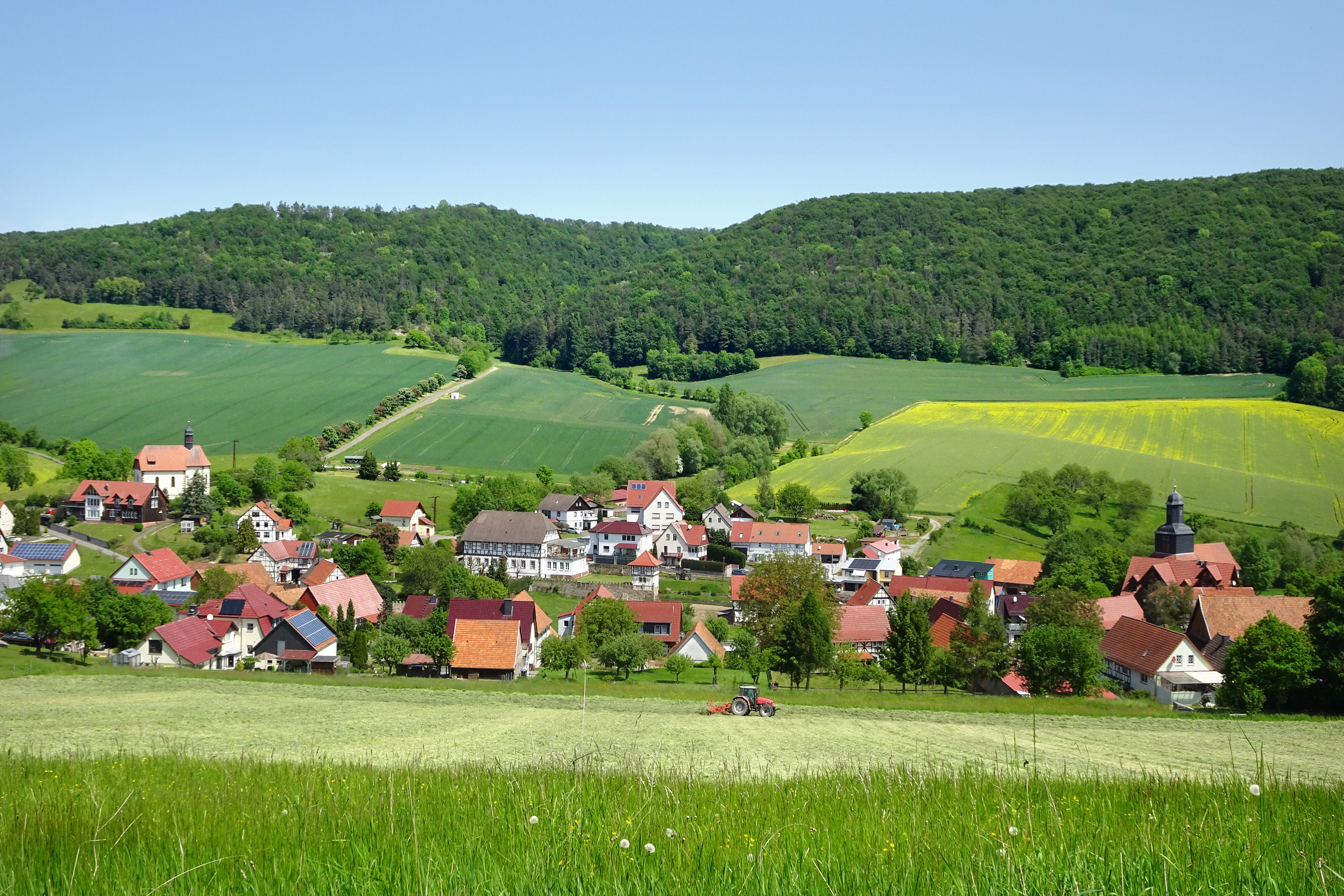 A wide, sunny landscape view of the village of Fretterode nestled in a green valley, with rolling hills and agricultural fields in the background.