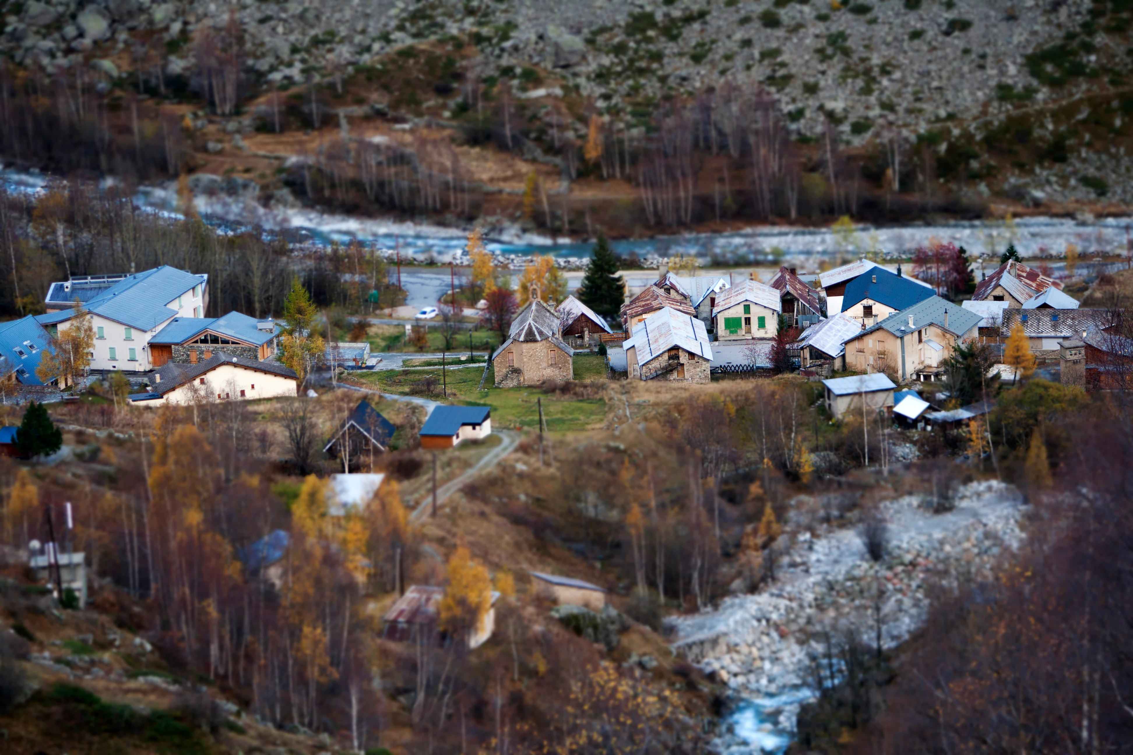 A high-angle view of the small village of La Bérarde, featuring stone buildings with grey roofs, set among golden autumn trees and rugged alpine terrain with a flowing river nearby.