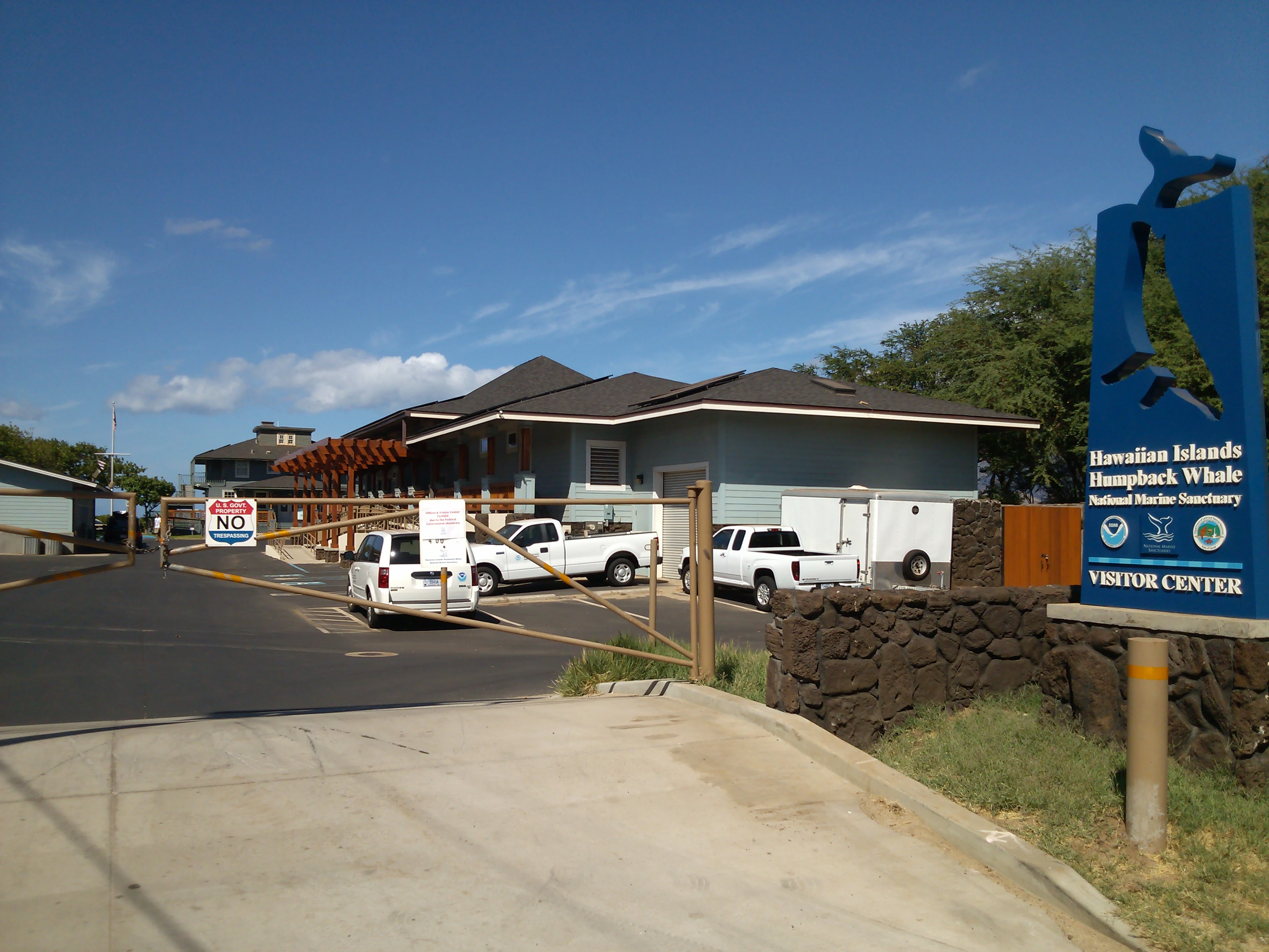 A view of the entrance to the Hawaiian Islands Humpback Whale National Marine Sanctuary visitor center, featuring a blue sign with a whale tail silhouette and a gated facility.