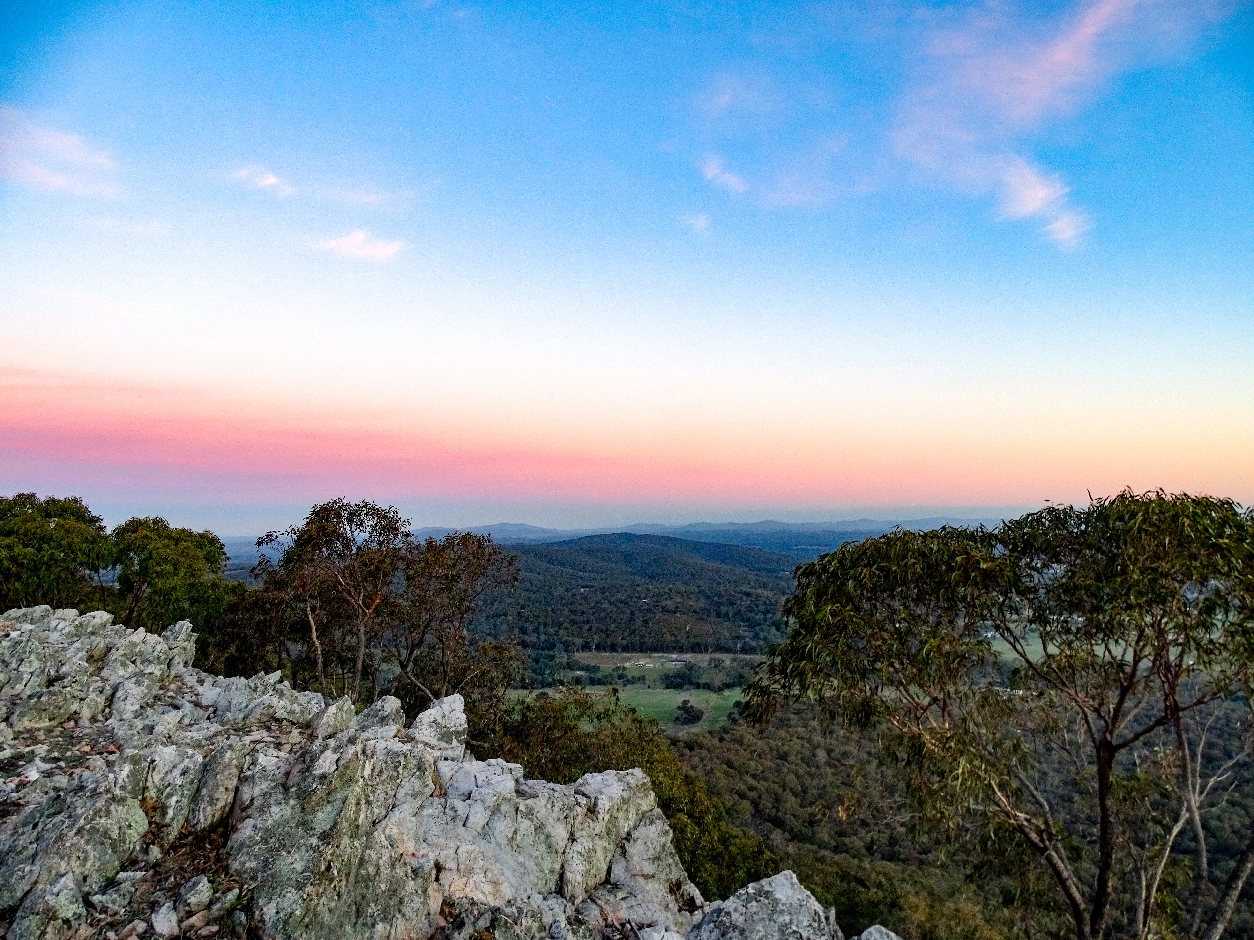 Rocky terrain in foreground, scattered trees, valley, and colorful sunset sky