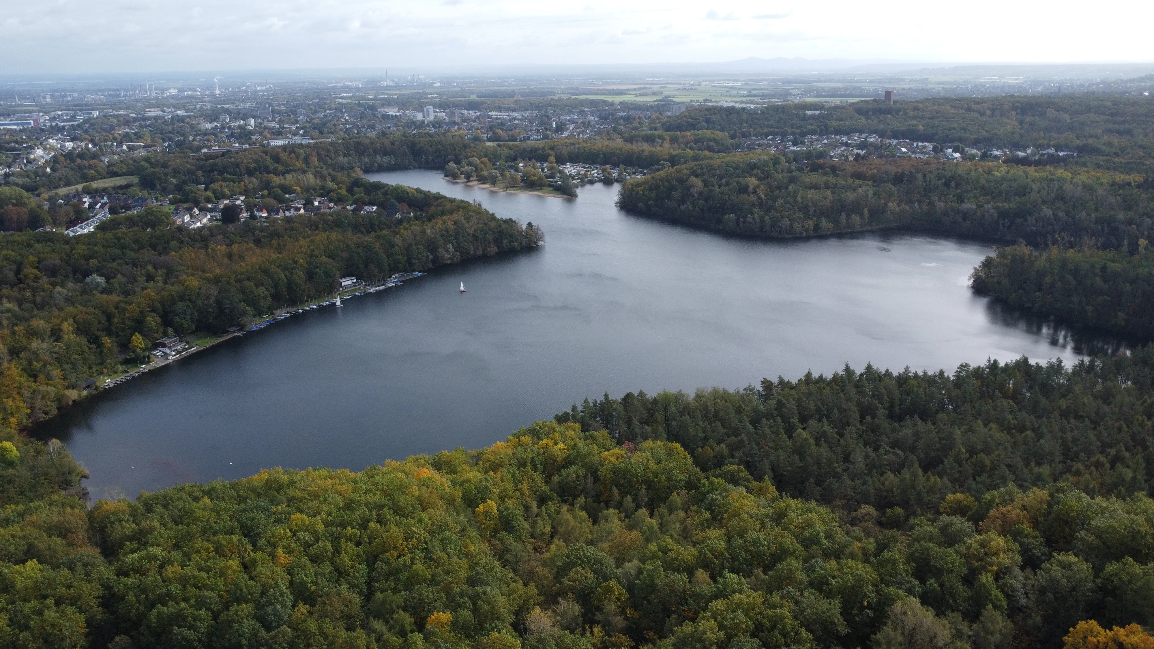 Aerial perspective of a large, dark lake surrounded by dense woodland and forest, with urban development visible in the distant background under an overcast sky.