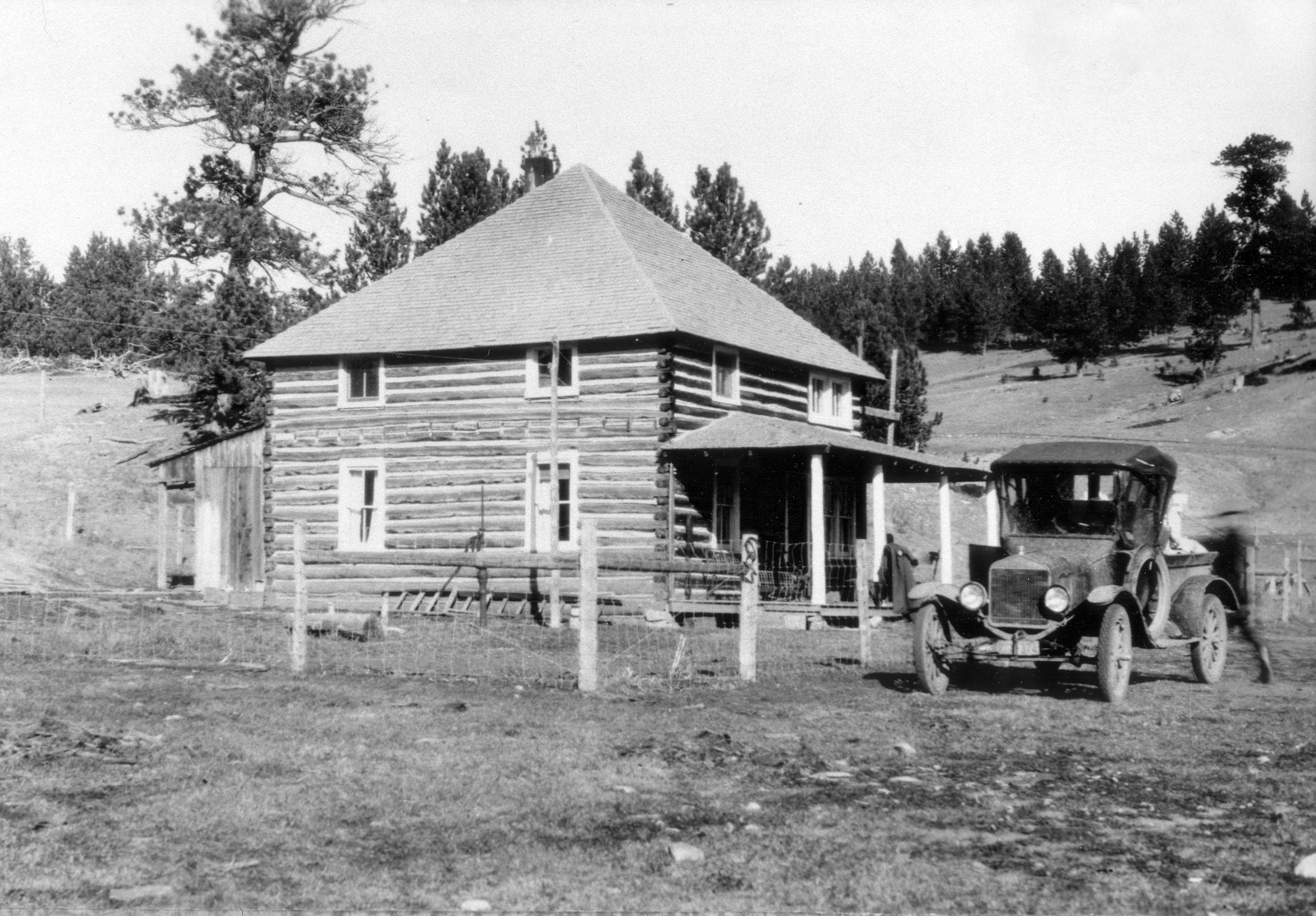 A black and white archival photograph of a two-story log cabin with a pyramid roof and a front porch, with a vintage automobile parked in front on a dirt landscape.