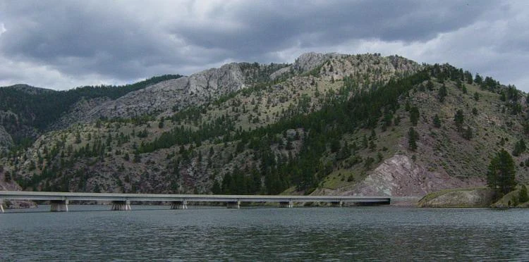 A long concrete bridge stretches across a wide river, backed by a large, tree-covered mountain slope under a cloudy sky.