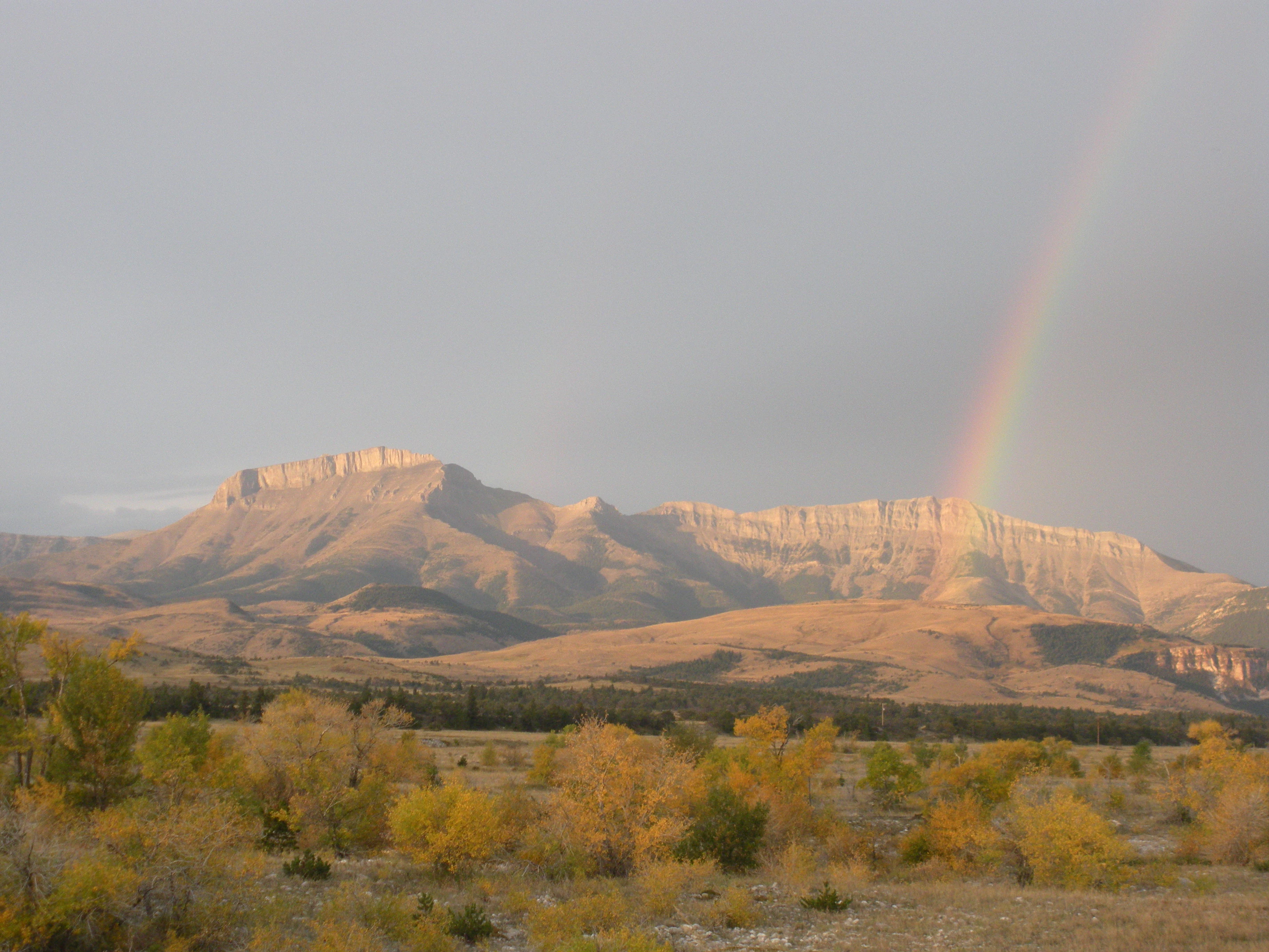 A wide landscape view of Ear Mountain with a rainbow stretching down toward the cliffs under a cloudy sky.