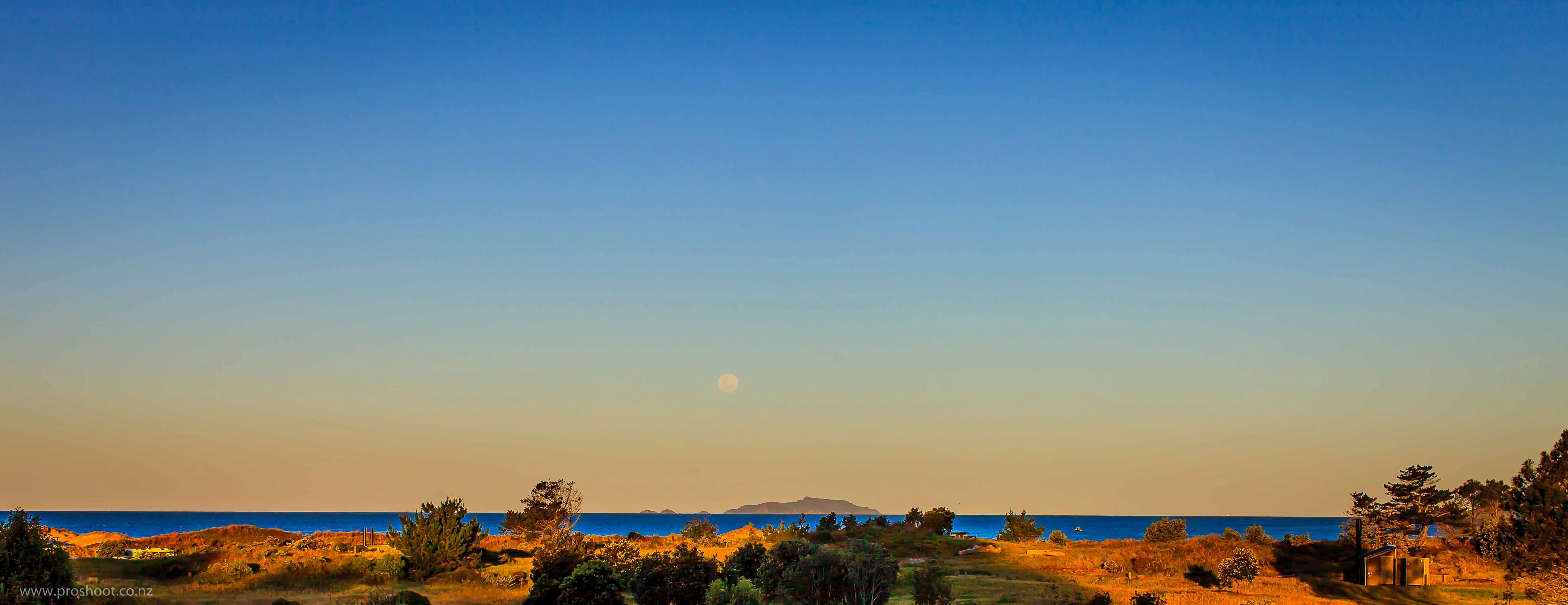Distant islands visible on the horizon across a body of water at sunset, with coastal scrub vegetation in the foreground.