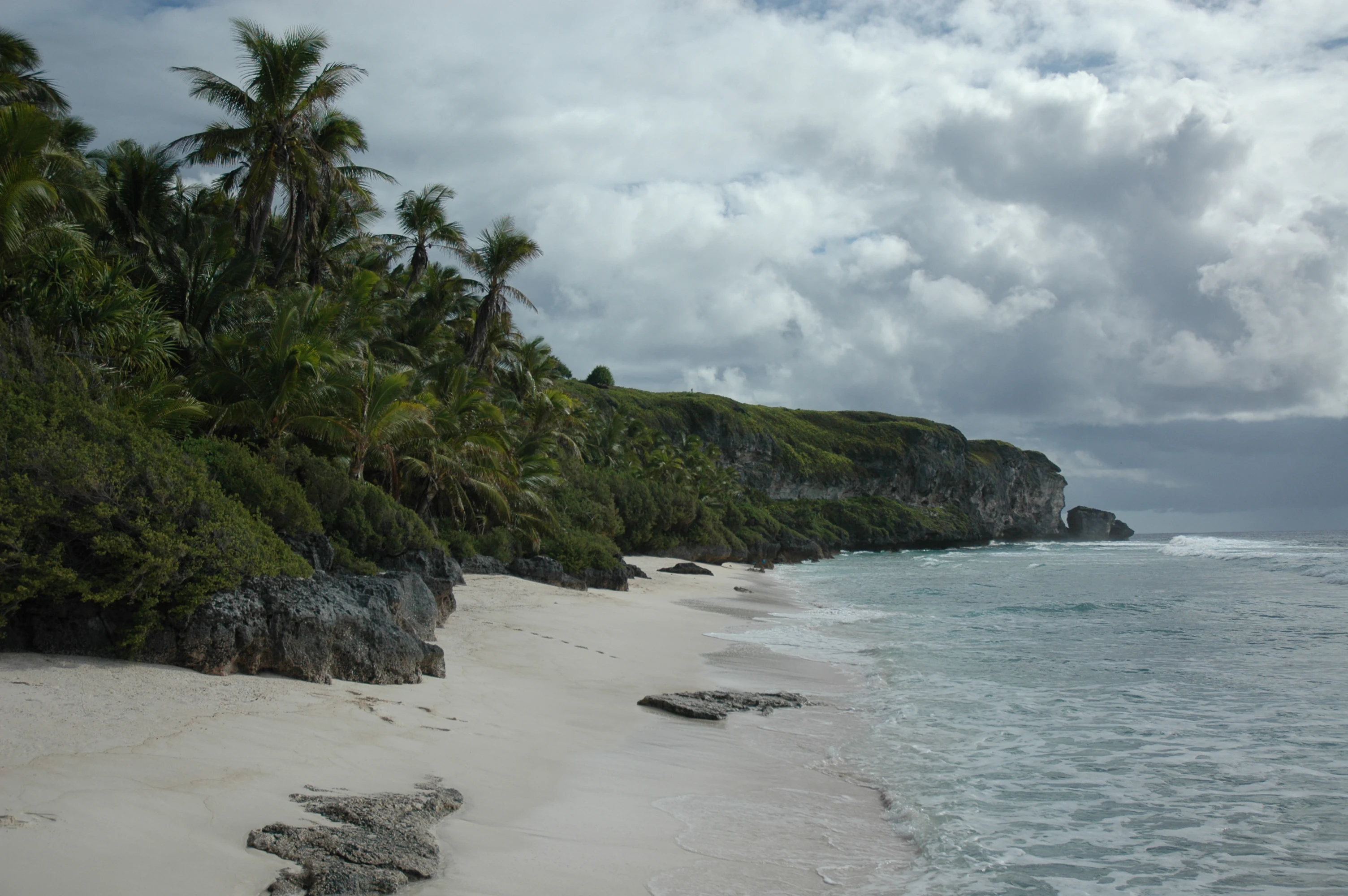A sandy beach stretches along a shoreline with rocky limestone cliffs topped with dense tropical vegetation, leading to the ocean under a cloudy sky.