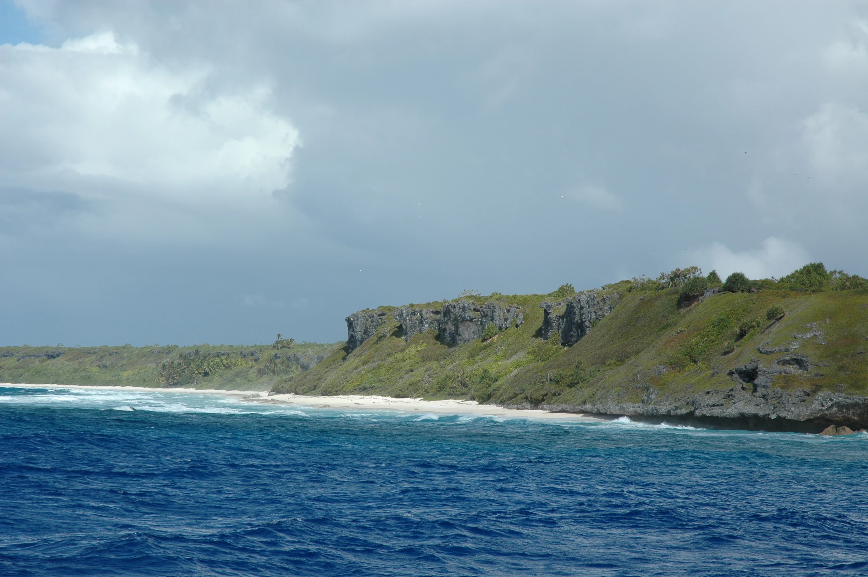A coastal view showing a white sand beach meeting turquoise waters, backed by raised coral cliffs covered in green vegetation under a cloudy sky.
